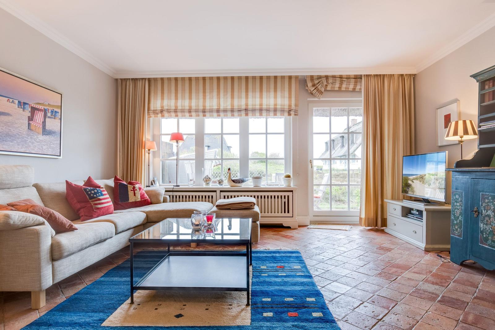 Wohnzimmer mit Sofa, Couchtisch und TV. Große Fenster mit Blick nach draußen.