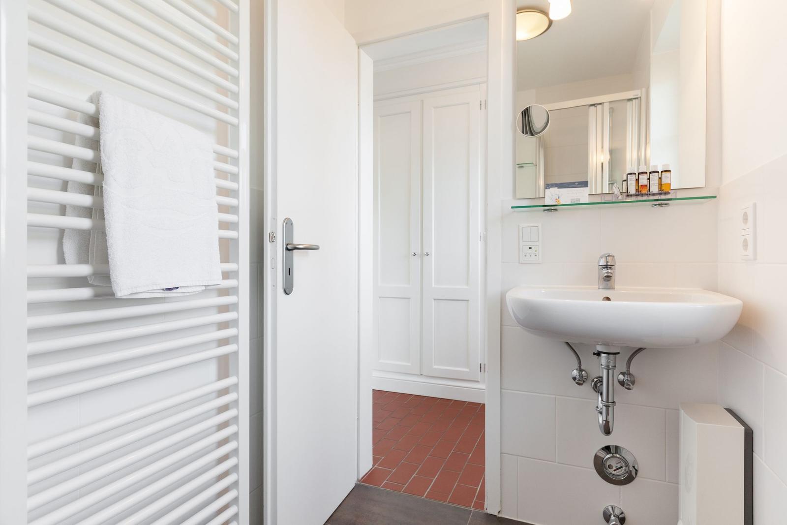Badezimmer mit Waschbecken, Spiegel und Handtuchhalter. Durchgang zur Garderobe mit rotem Boden.