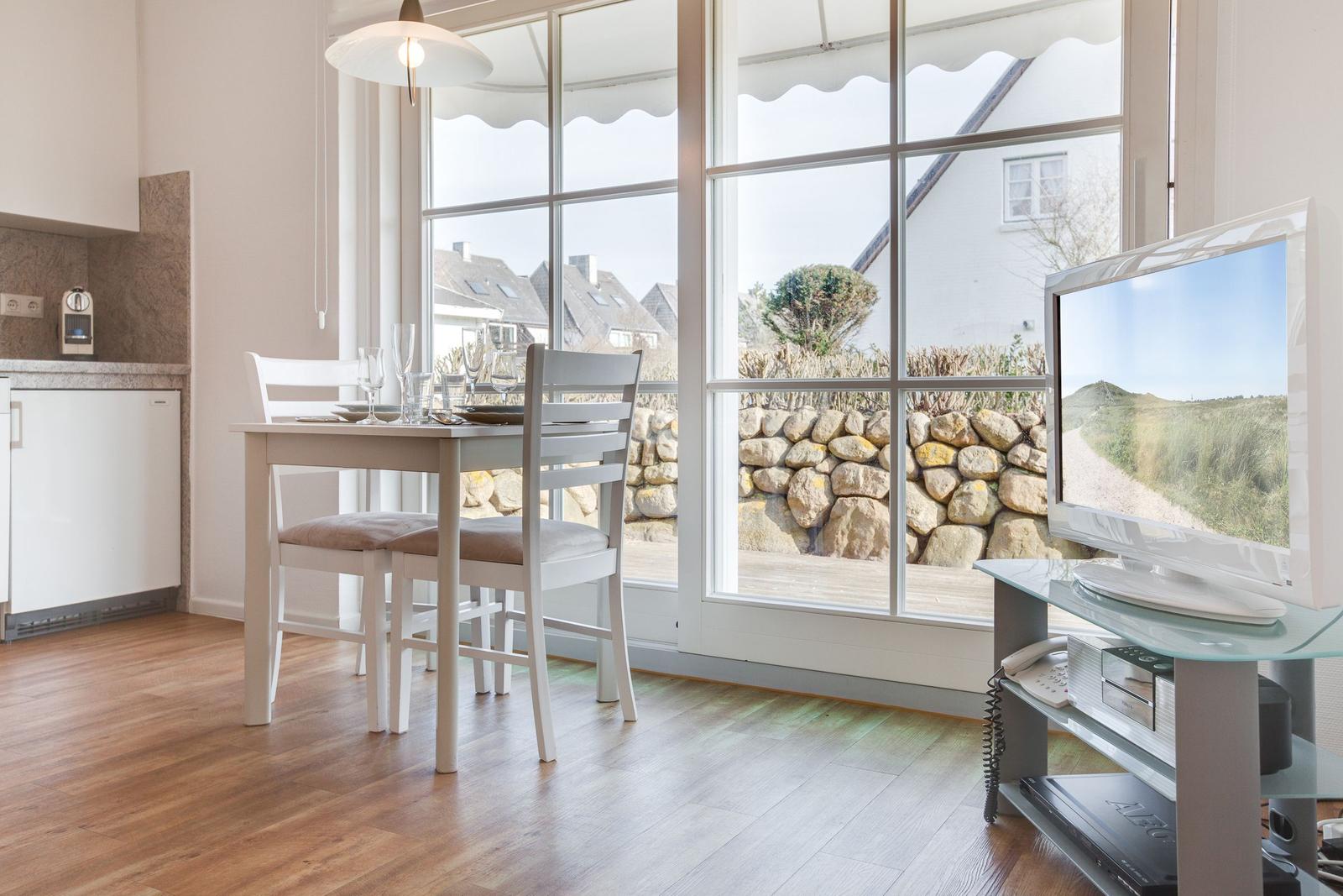 Essbereich mit Tisch, Stühlen und Fernseher. Große Fenster mit Blick auf Steinmauer und Garten.