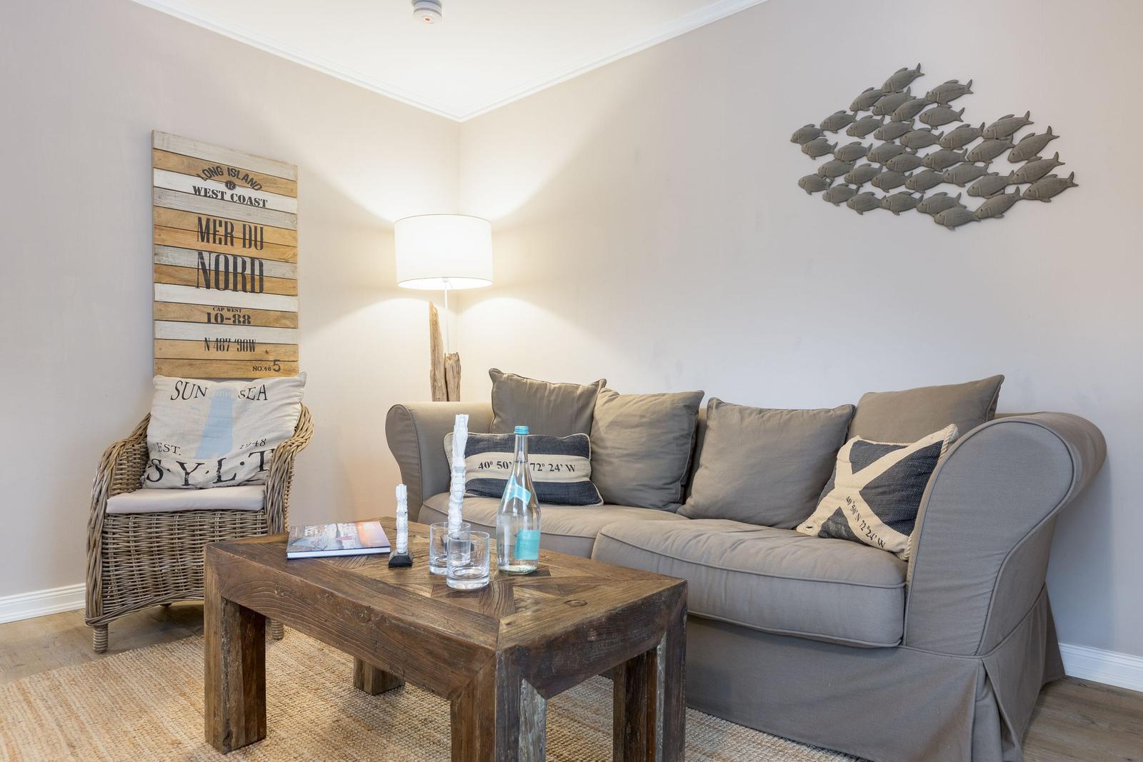 Living room with sofa, armchair, and wooden table. Wall decor with fish motif and wooden sign.