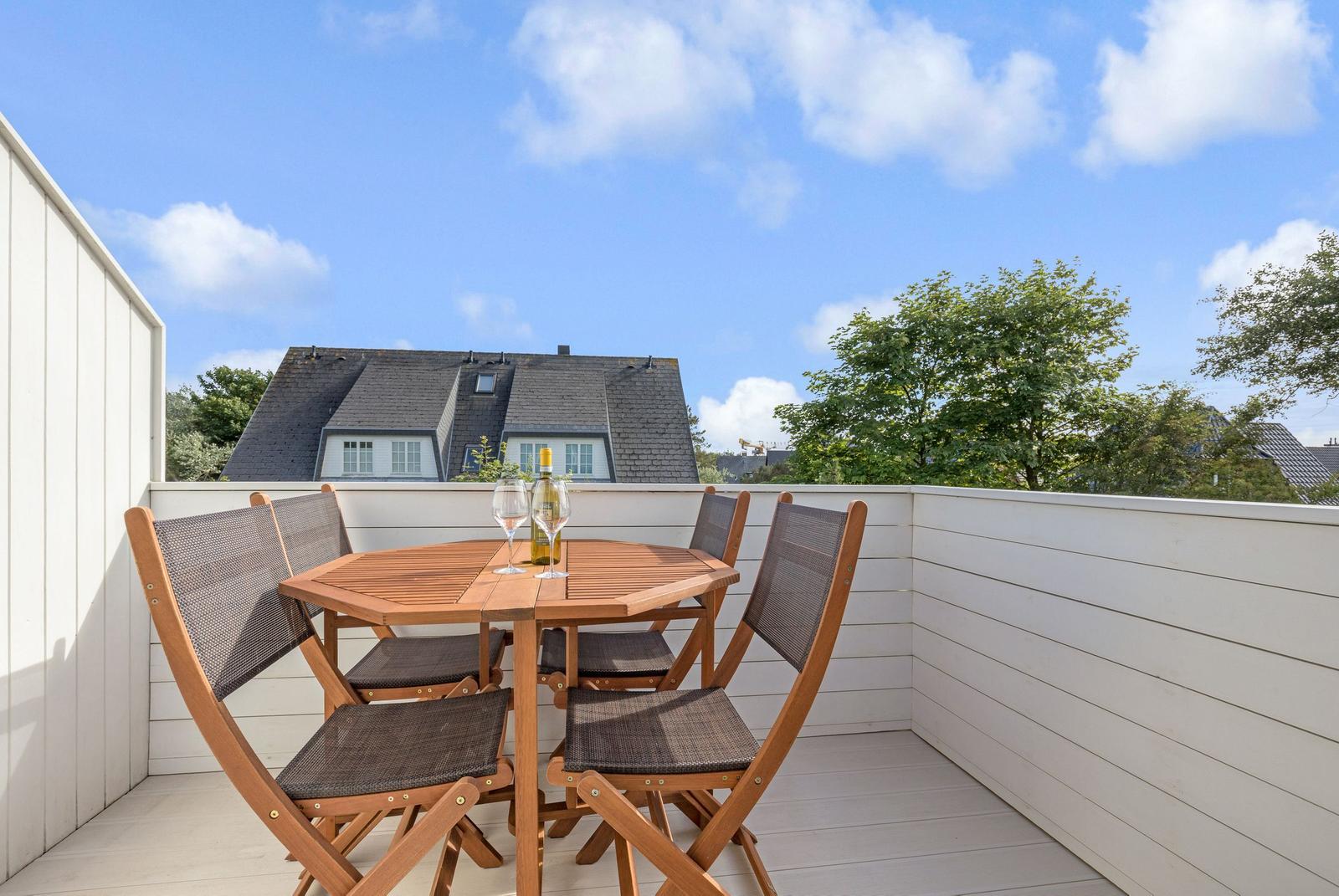 Terrasse mit Holztisch und vier Stühlen. Im Hintergrund sind Häuser und Bäume zu sehen.