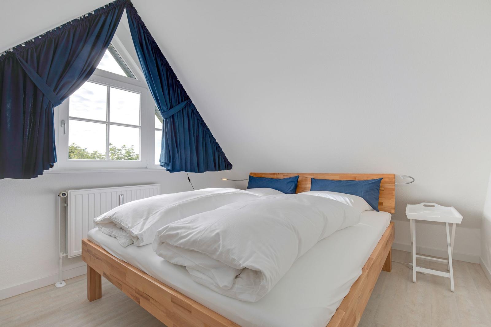 Schlafzimmer mit Holzbett, blauen Vorhängen und Fenster. Neben dem Bett steht ein weißer Nachttisch.