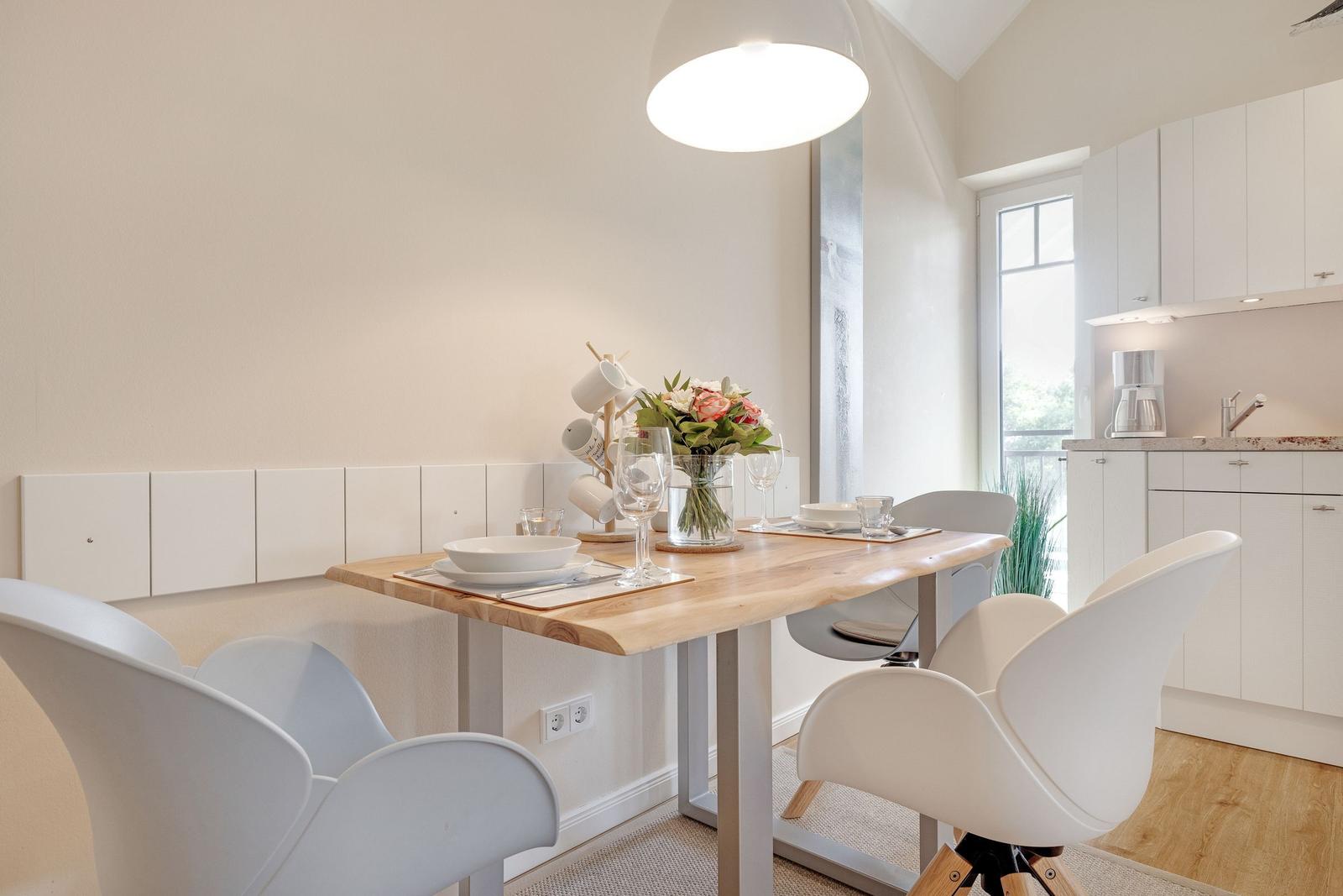 Dining area with wooden table and white chairs. Kitchen with white cabinetry and window.