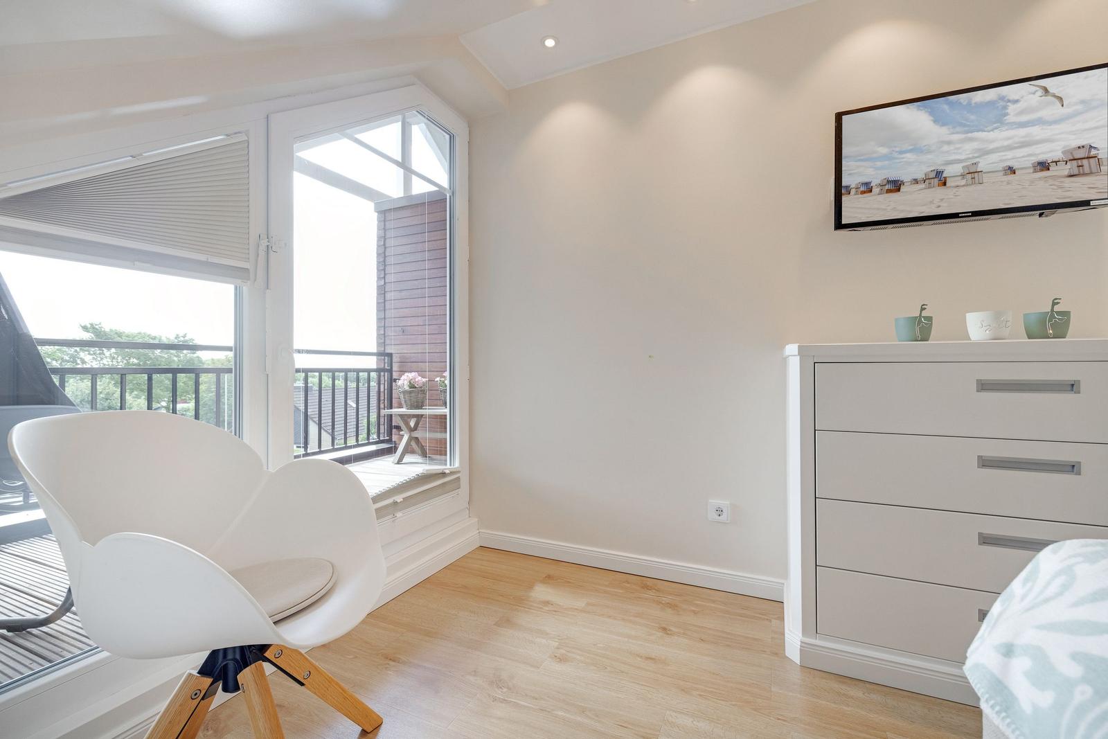 A bedroom with a white chair, dresser, and TV on the wall. Balcony with view of outdoor area.