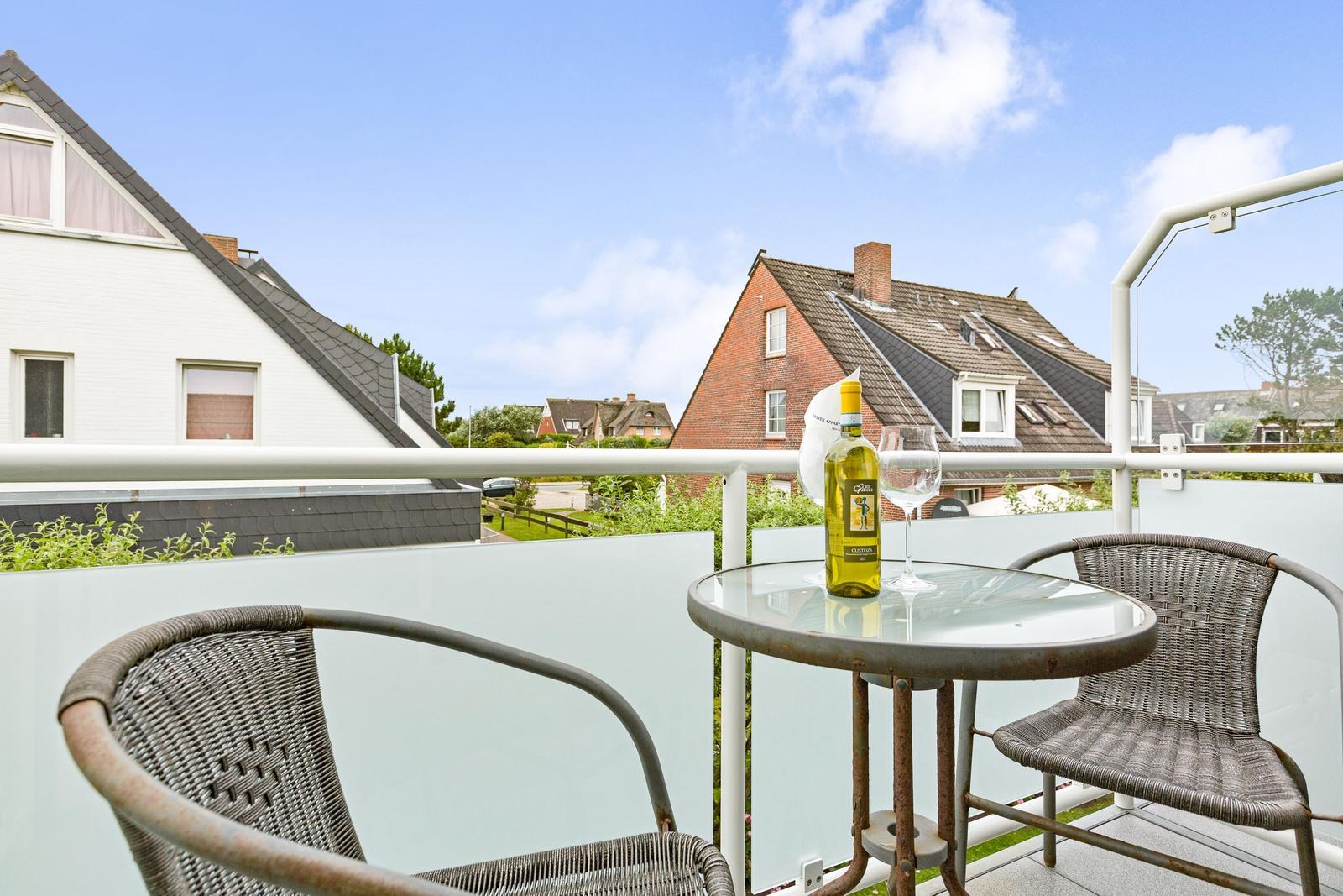 Terrasse mit Tisch, zwei Stühlen und Weinflasche. Blick auf Häuser und Grünflächen.