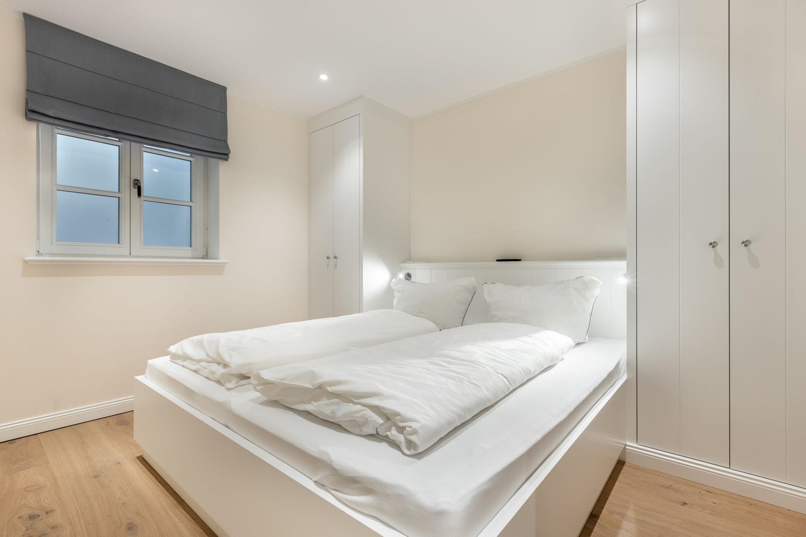 Schlafzimmer mit weißem Bett, Kleiderschrank und Fenster mit Rollladen.