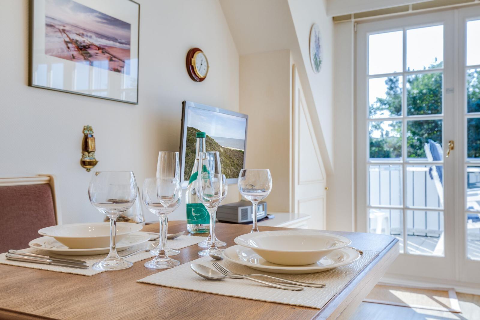 Dining table set with dishes, glasses, and bottle. Windows open to balcony with green view.