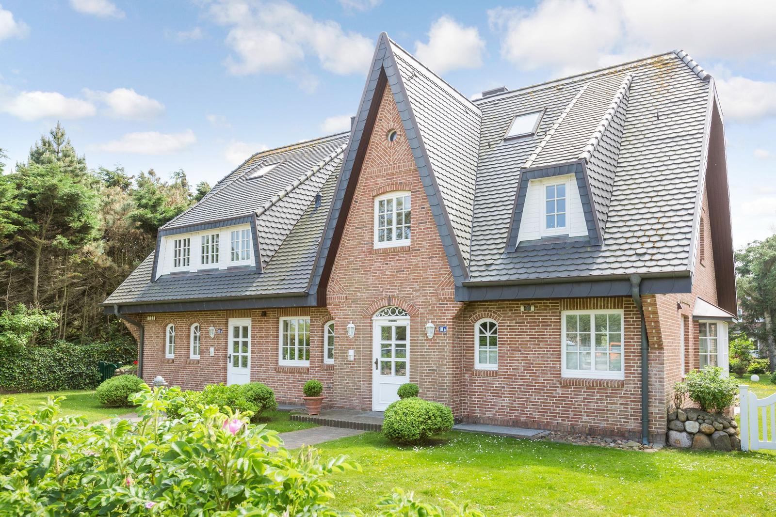 Red brick house with dormer windows and white entrance. Green lawn and trees surround the house.