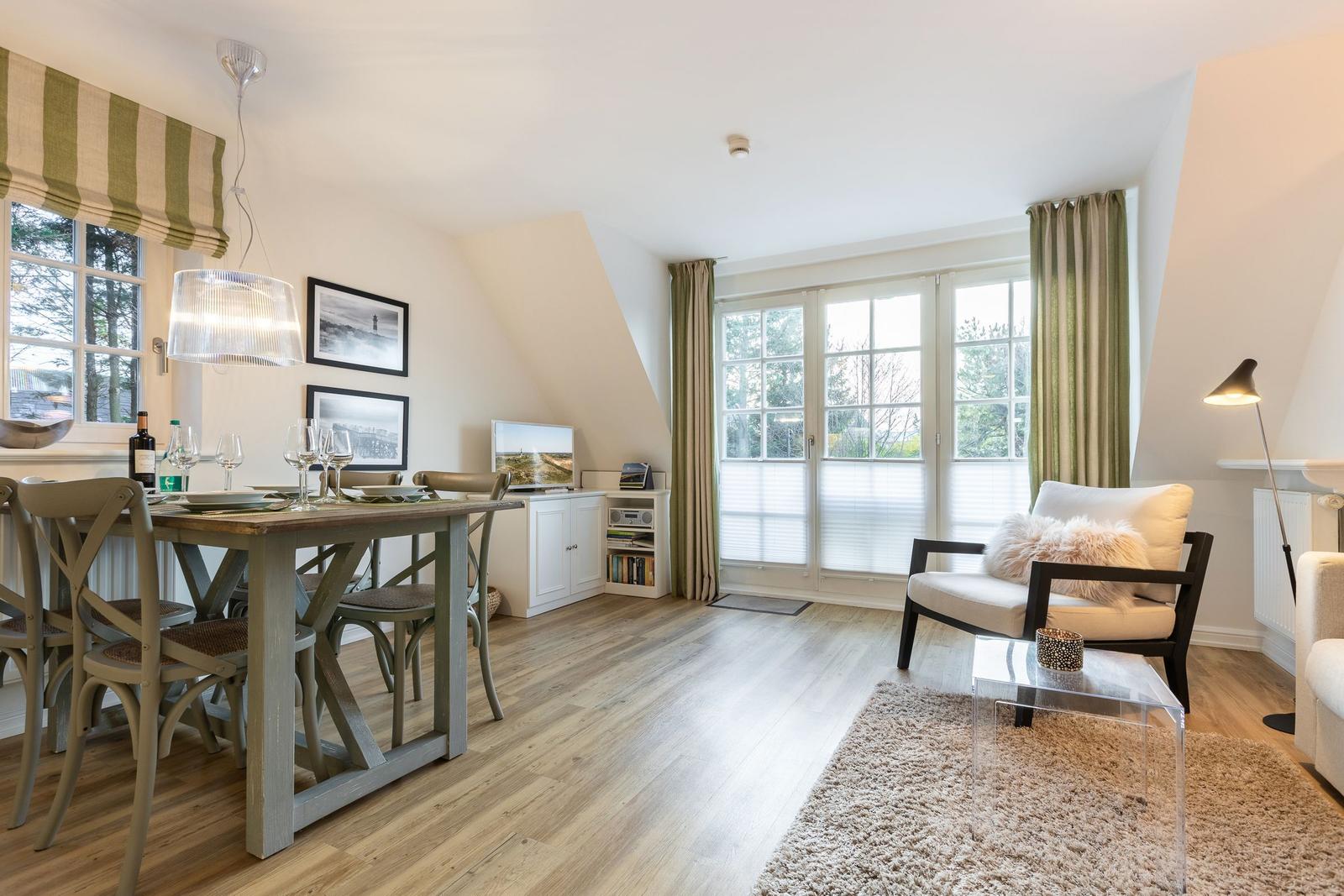 Dining area with table and chairs, living area with armchair and sofa, wooden floor, large windows.
