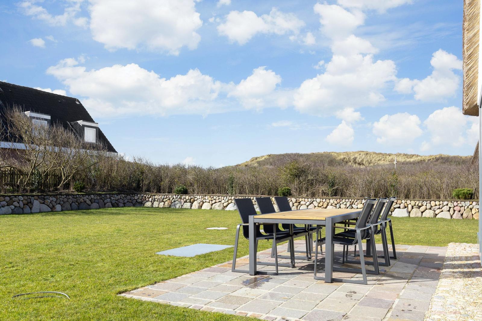 Hinterhof mit Tisch und Stühlen, Grasfläche und Steinmauer. Blick auf Dünen im Hintergrund.