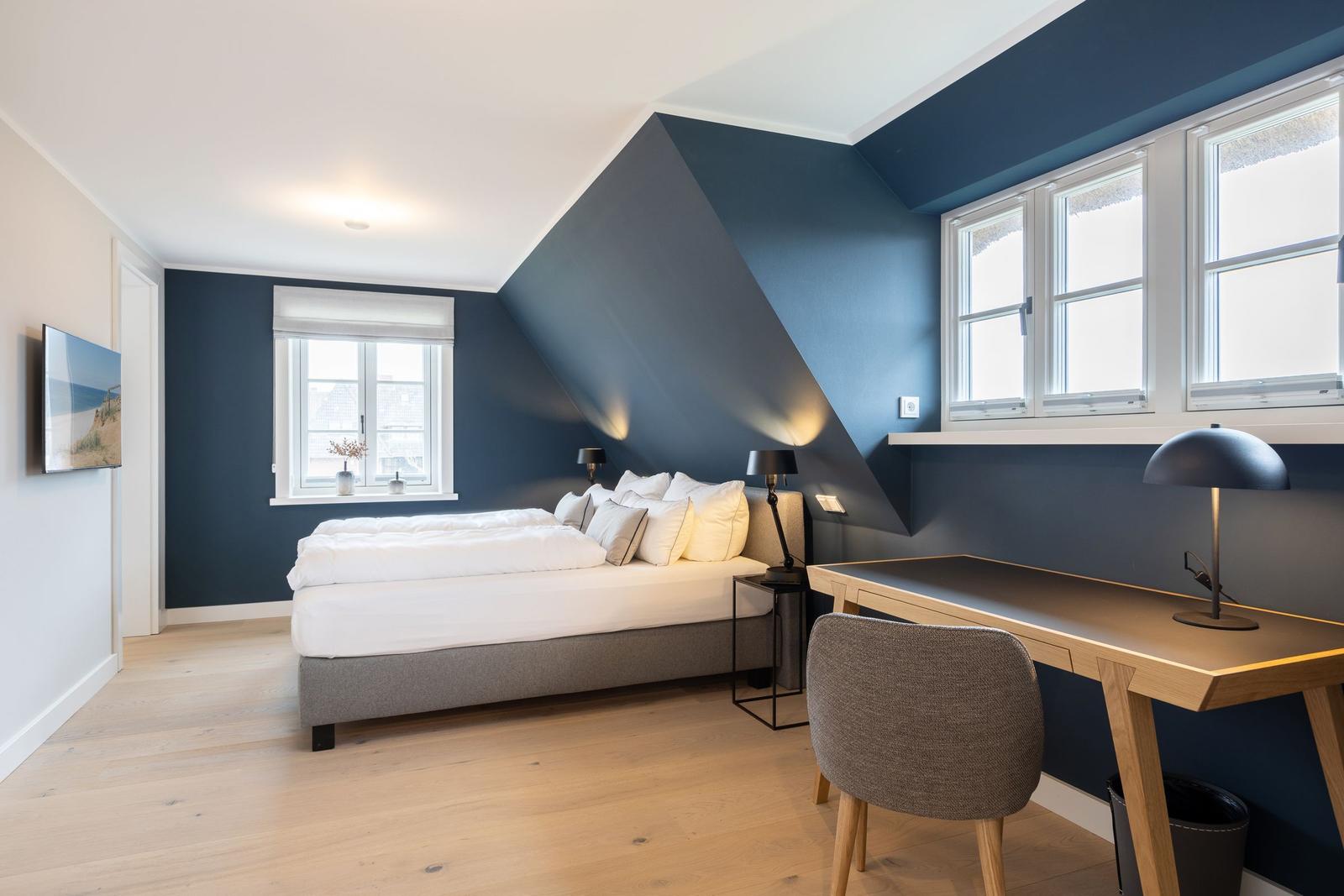 Schlafzimmer mit Bett, Schreibtisch und Fenstern.