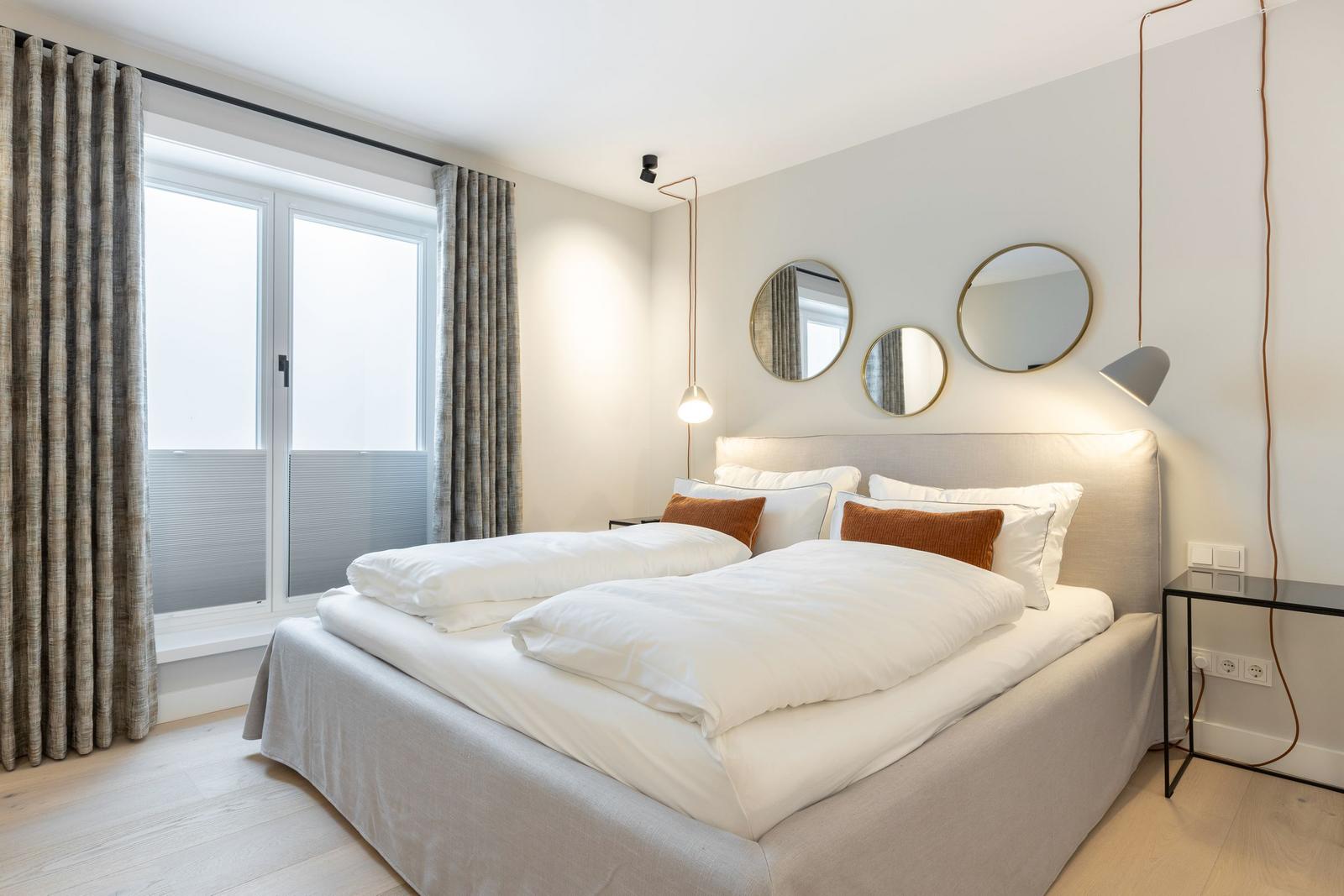 Schlafzimmer mit Doppelbett, runden Spiegeln und Fenster mit Vorhängen.