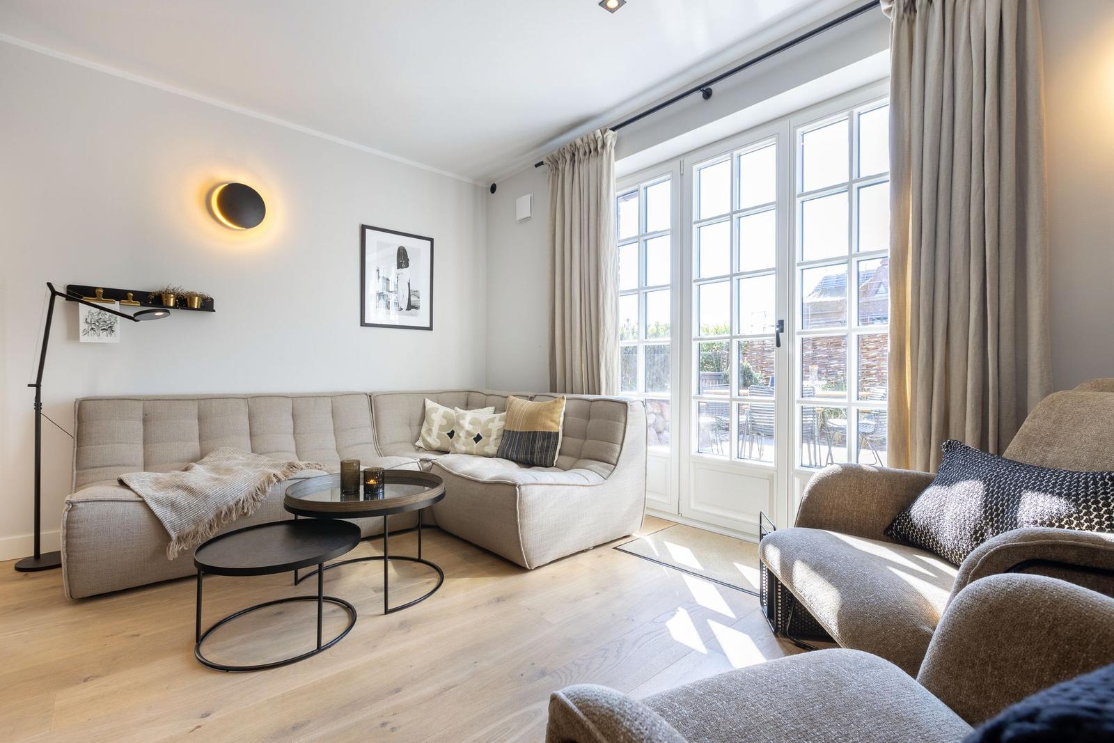 Living room with sofa, armchairs, and tables. Large windows and wooden floor.