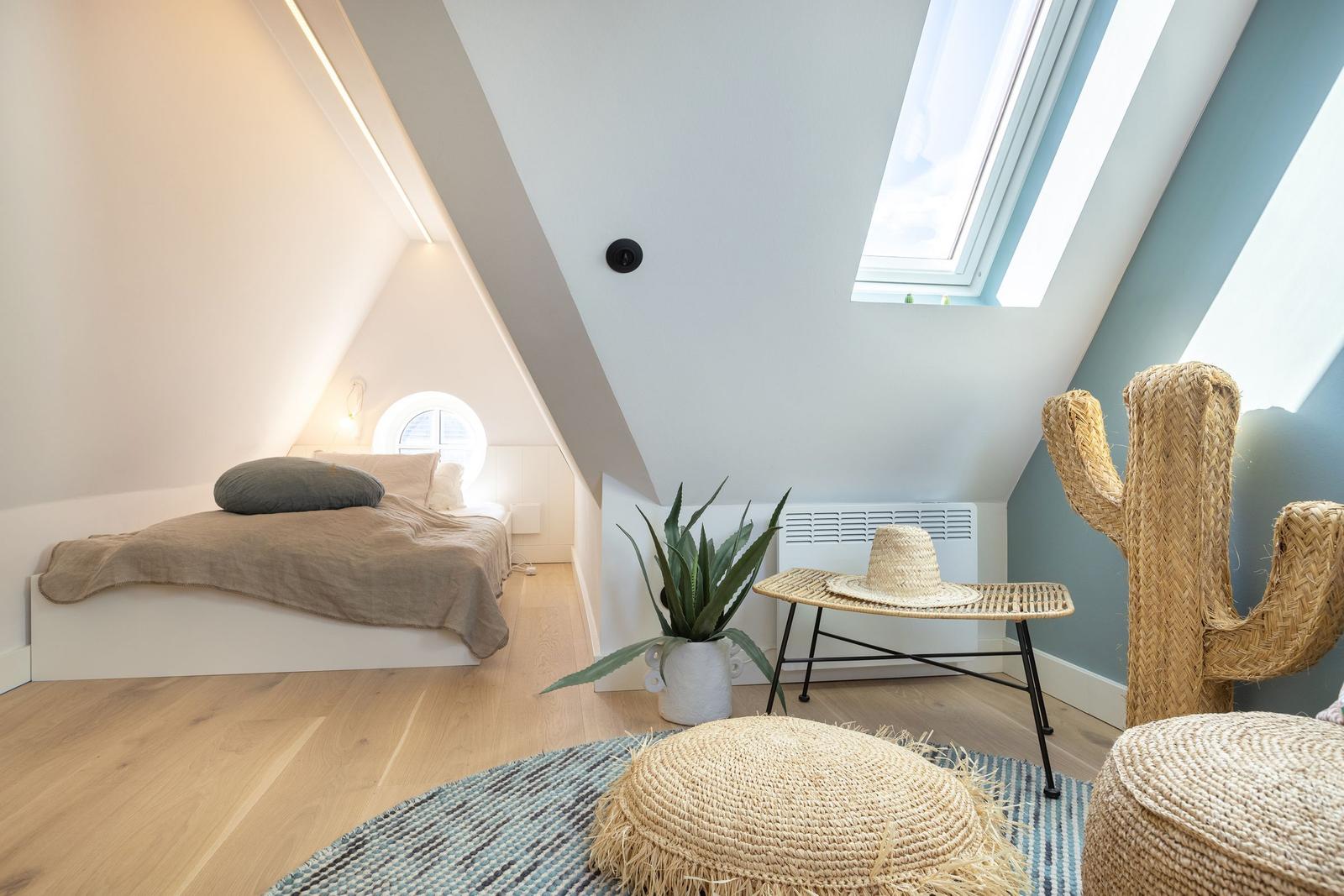 Bedroom with sloped ceiling, bed, plant, and seating under skylight.