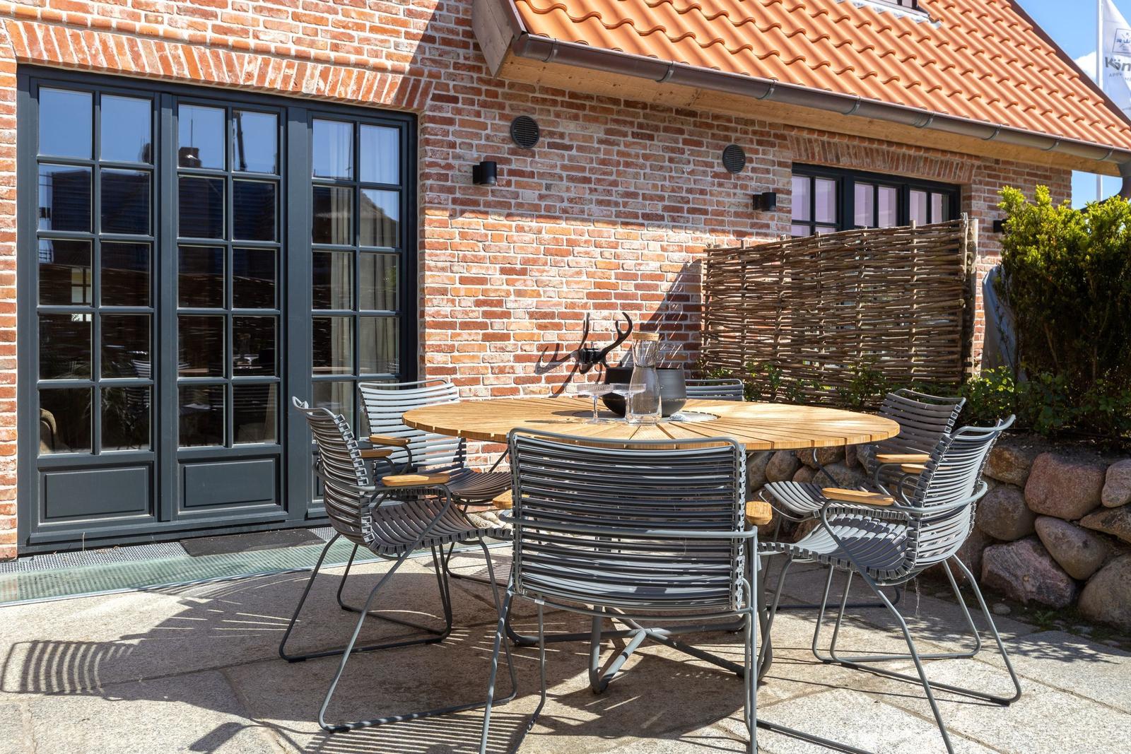 Terrace with round table and chairs in front of brick house with windows and roof.