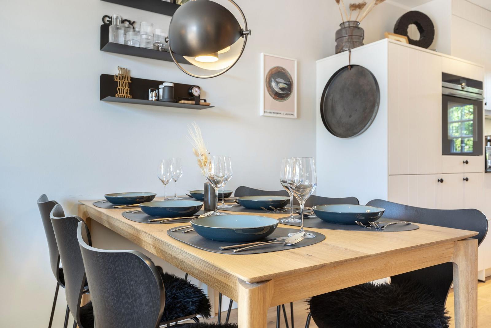 Dining table with six chairs and tableware. Background: kitchen cabinets and wall shelves.