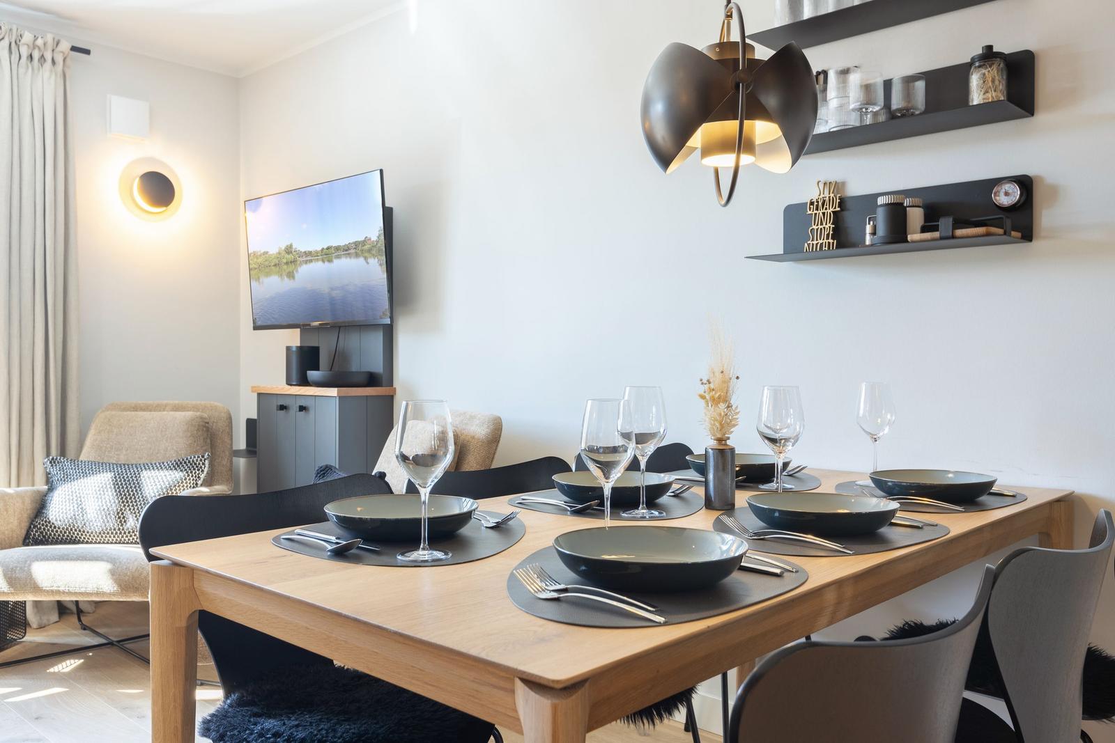 Dining table with six chairs and cutlery. Wall shelves and TV in the background.