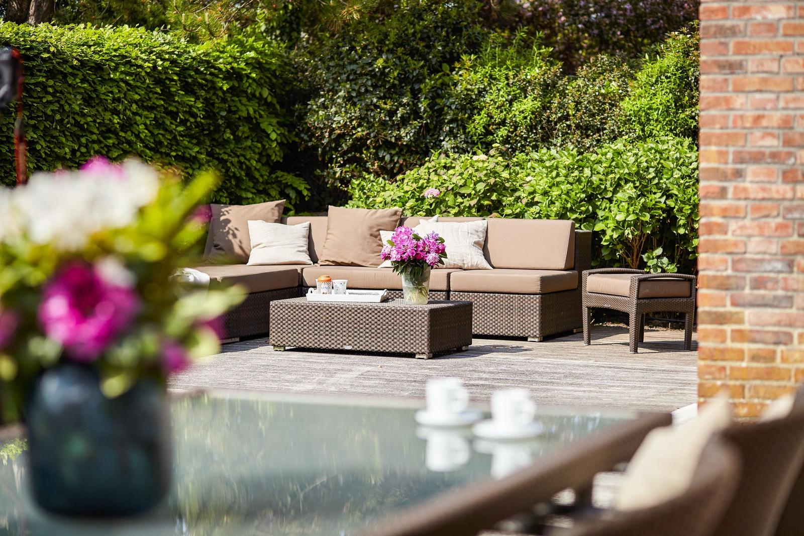 Terrace with seating, flowers, and glass table. Background: green hedge and brick wall.