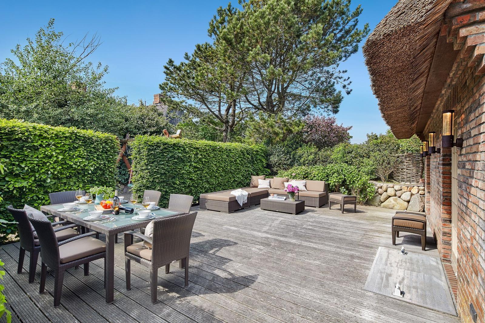 Terrace with seating area, dining table, and garden. Wooden deck, hedge, and tree visible.