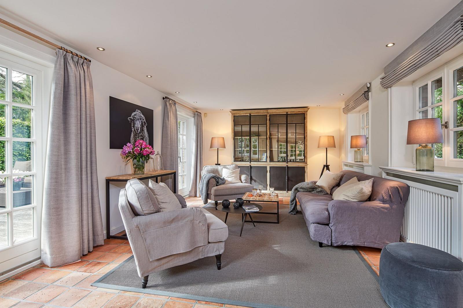 Living room with sofa, armchair, and table. Windows, curtains, and wall art are visible.