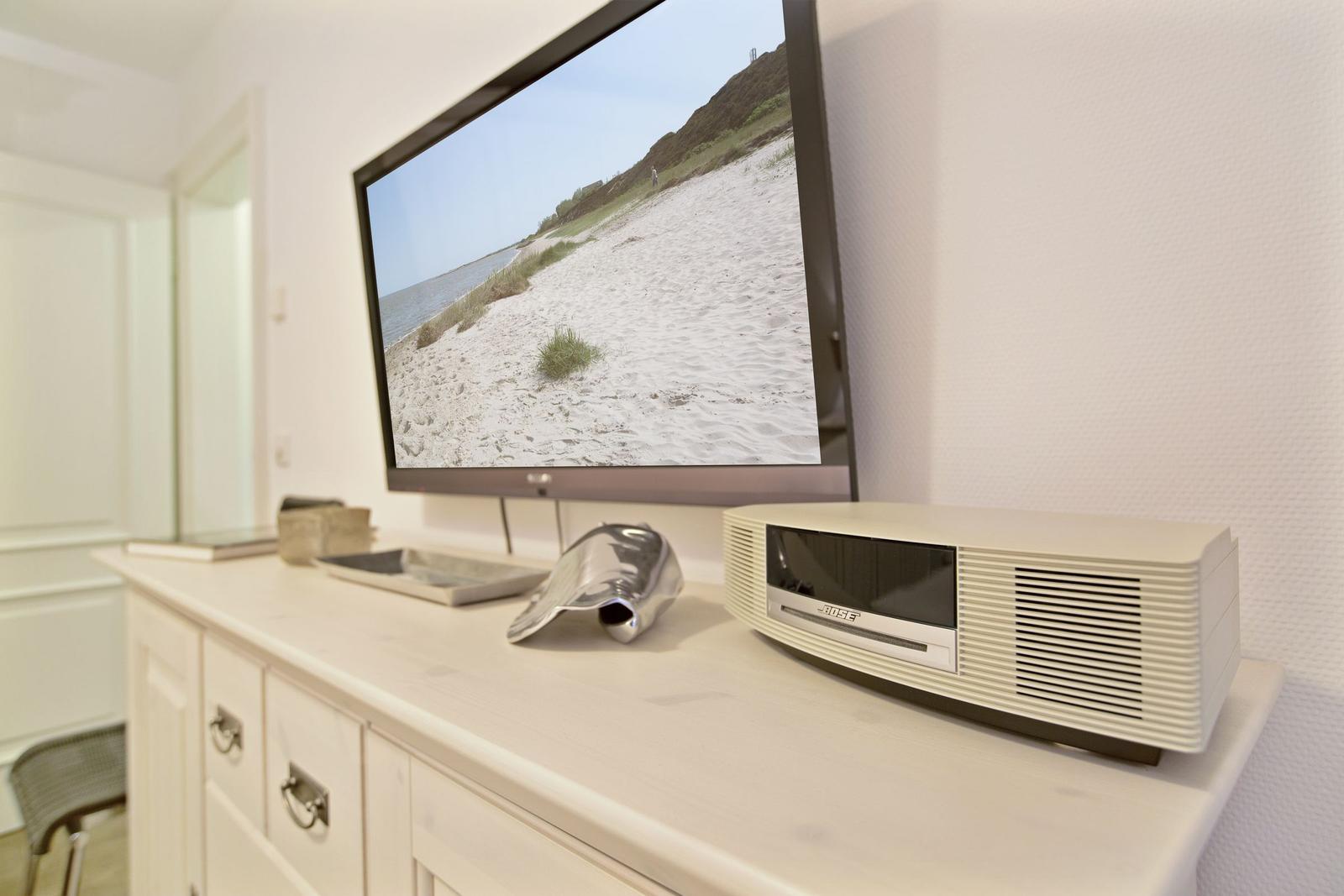TV auf Sideboard zeigt Strandbild, daneben steht ein weißer Lautsprecher.