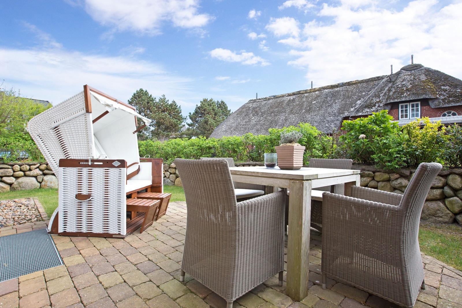 Terrasse mit Esstisch, Stühlen und Strandkorb. Hintergrund: Haus mit Strohdach.