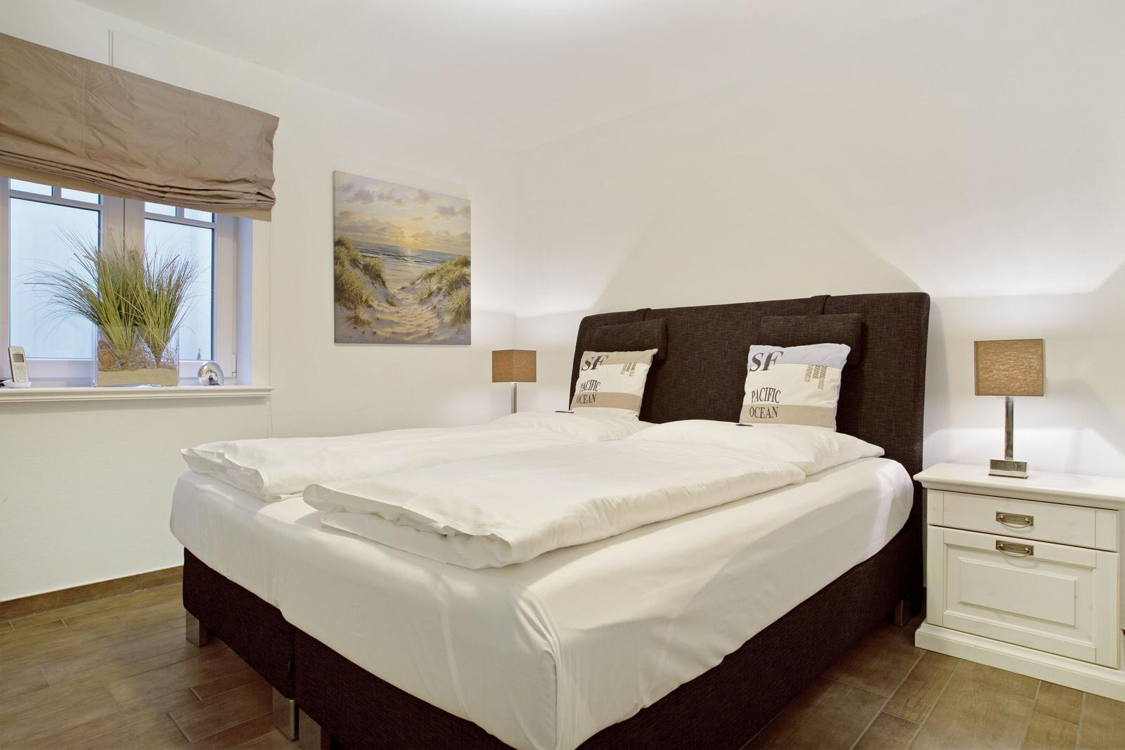 Schlafzimmer mit Bett, Nachttisch und Fenster. Wandbild zeigt Strand.