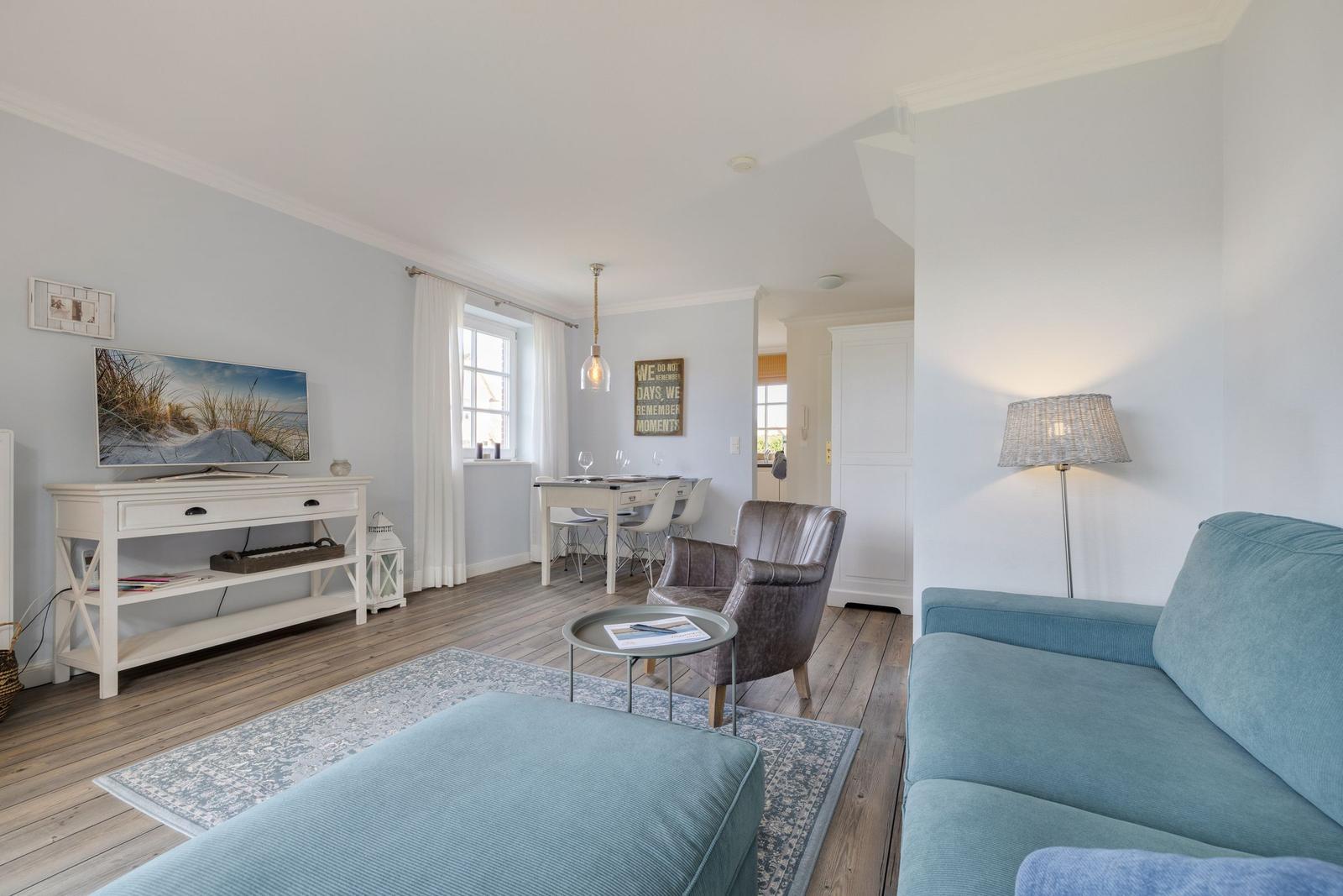 Living and dining area with sofa, armchair, and TV. Wooden floor and light walls.