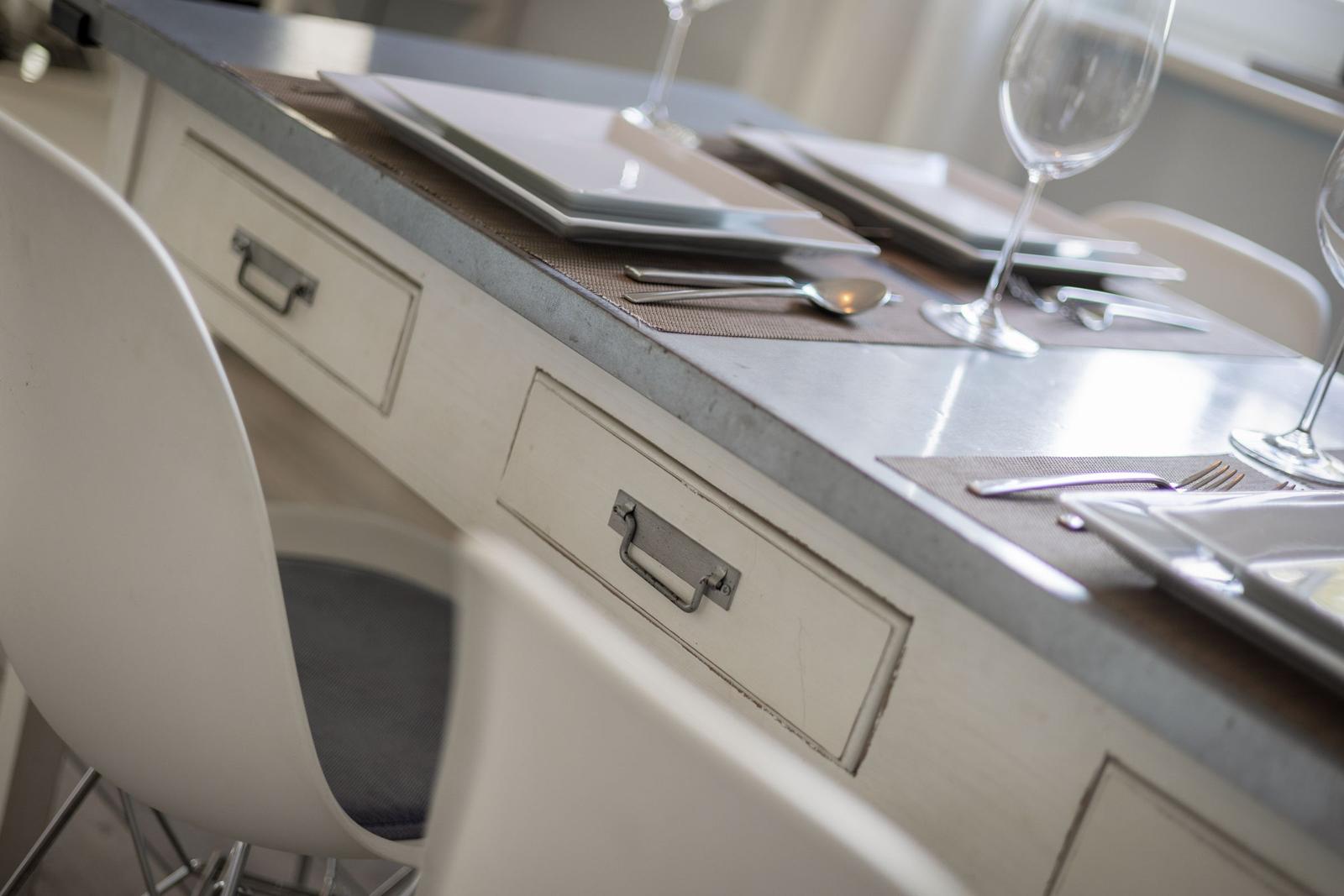 Dining table with chairs, cutlery, and glasses. Wooden surface with drawers.