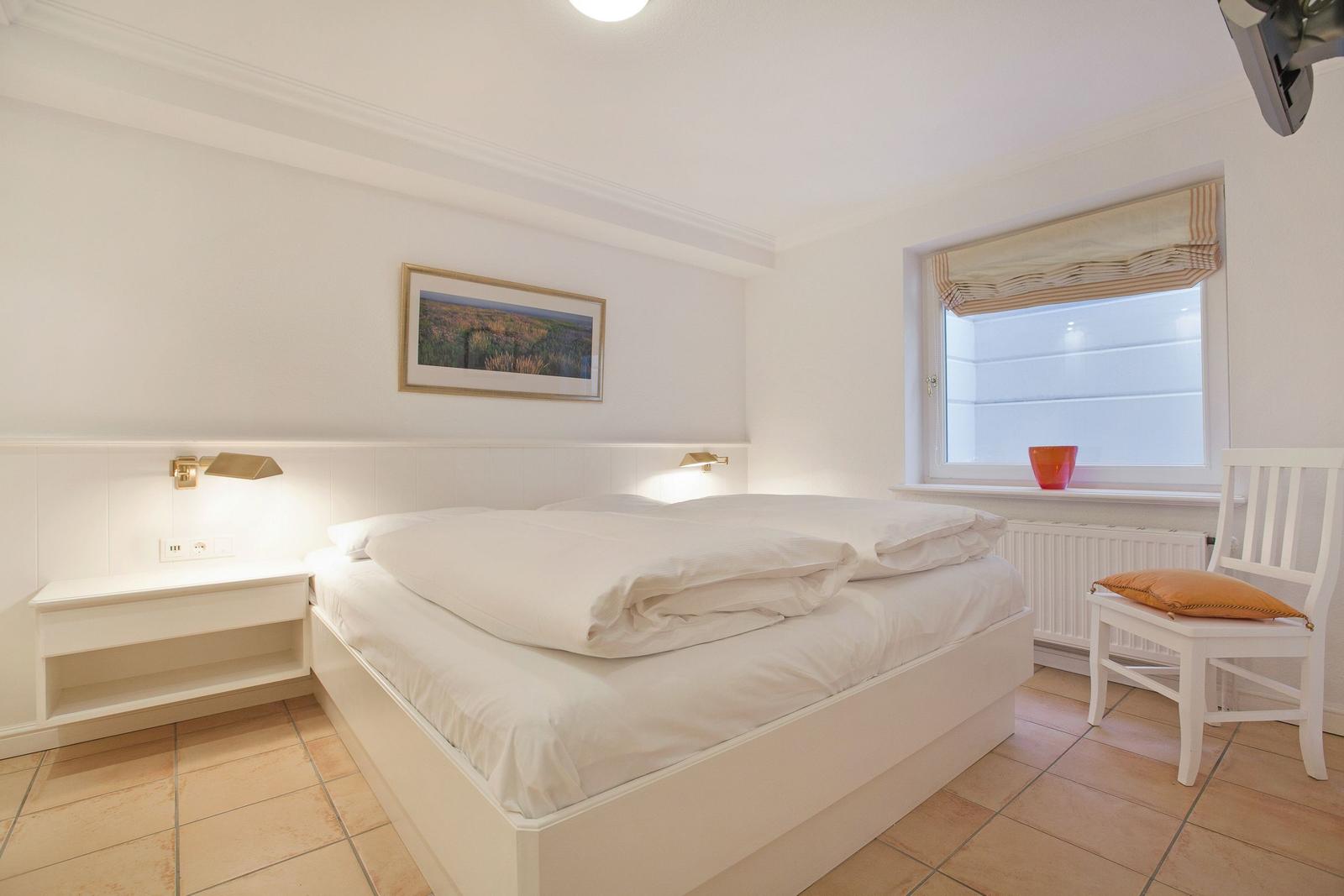 Bedroom with white bed, nightstand, and chair beside window.