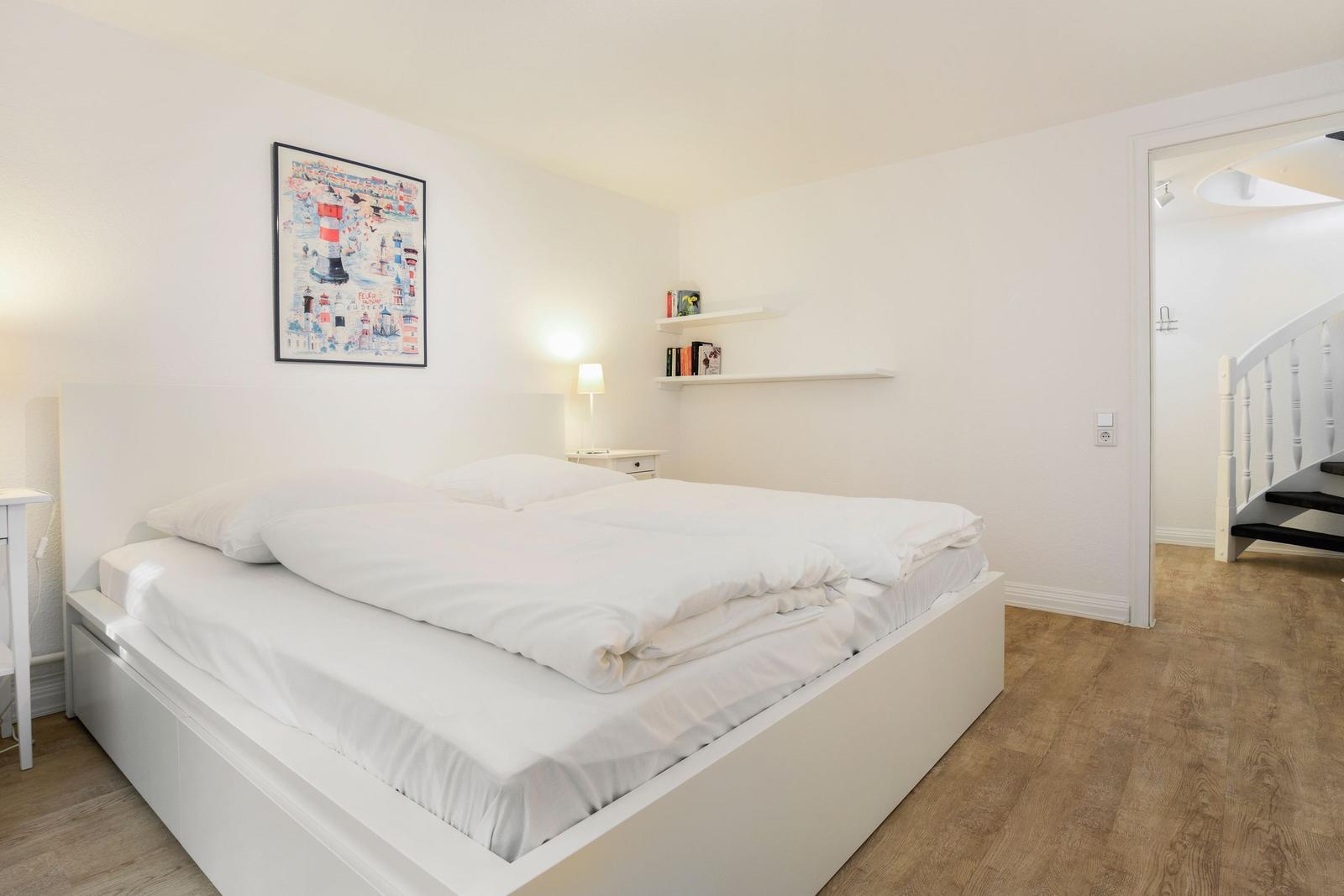 Bedroom with white bed, wooden floor, and wall art. Door leads to staircase.