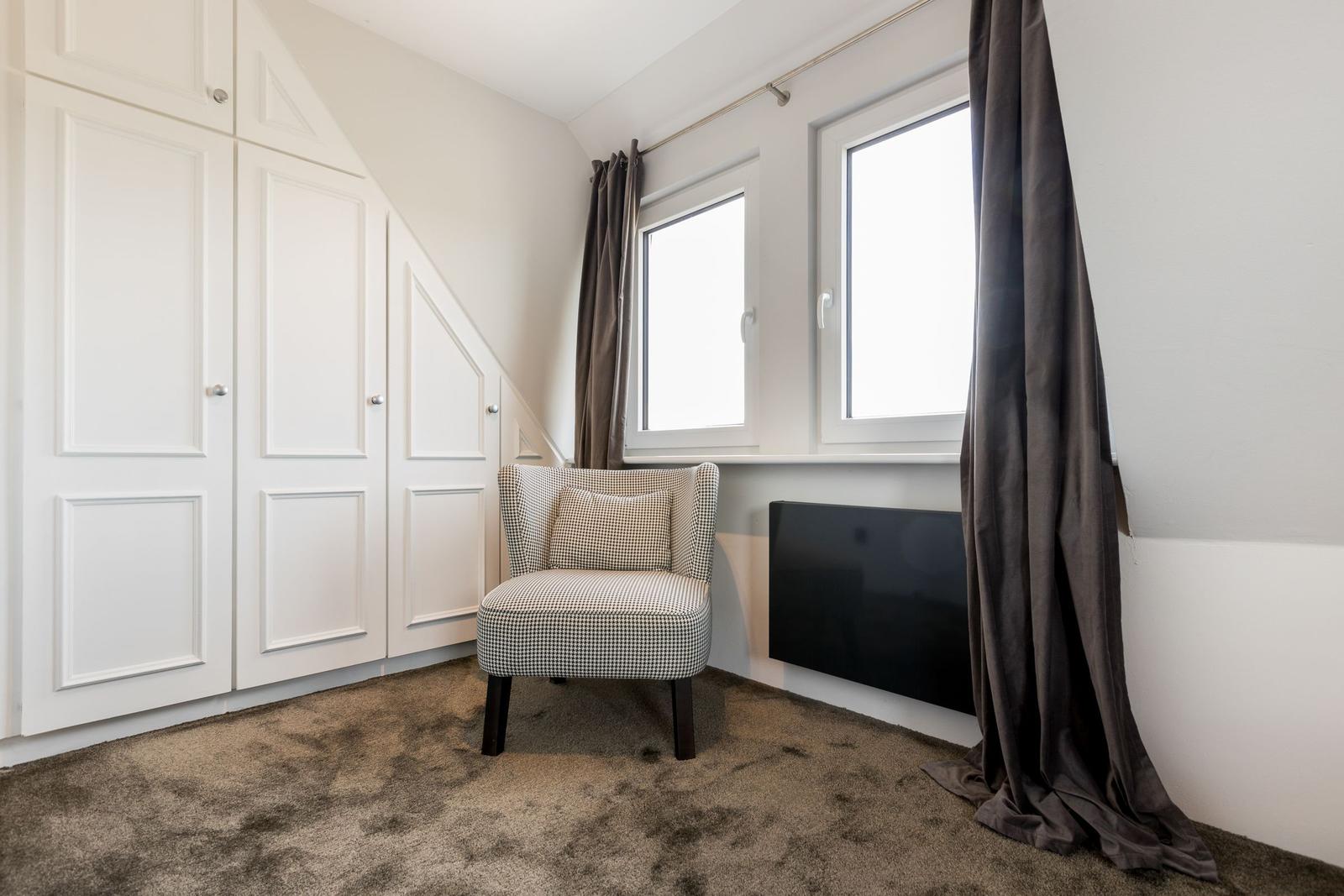 Zimmer mit weißen Schränken, Sessel und Fenstern mit Vorhängen.