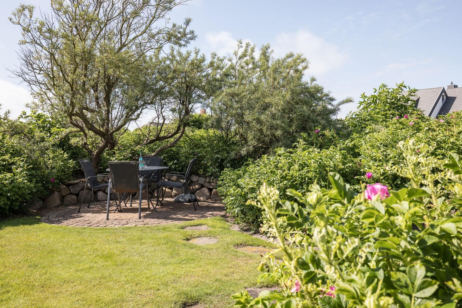 Garden with table, chairs, and blooming rose hedges under trees.