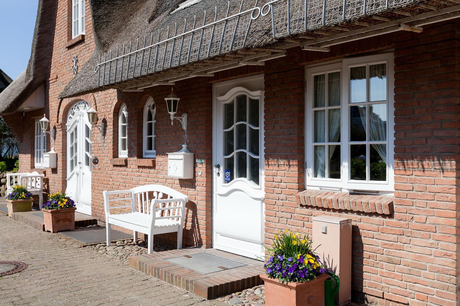 Red brick house with thatched roof, white doors and windows, flower pots and bench at entrance.