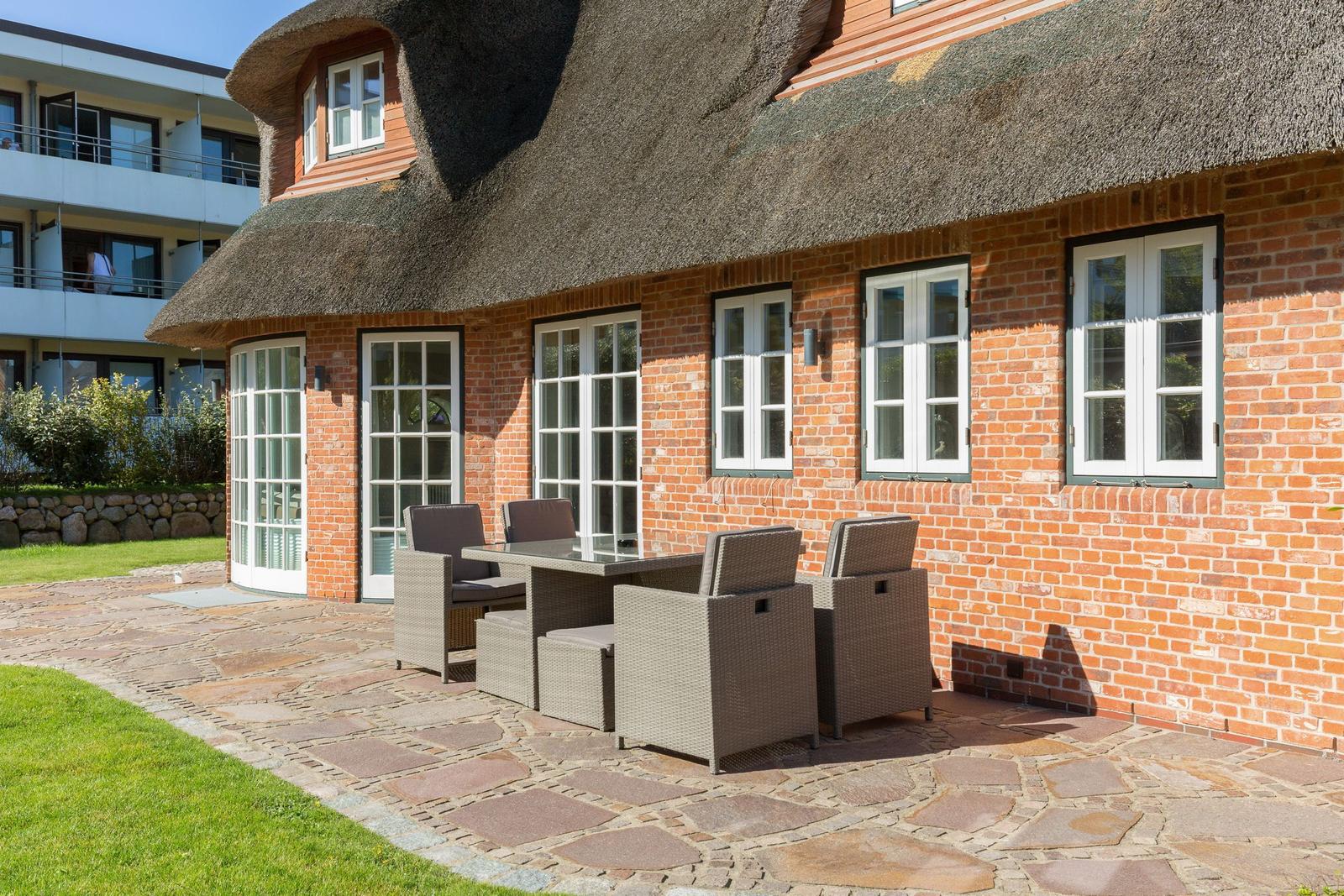 Rostrote Ziegelmauer mit strohgedecktem Dach und Terrasse mit Sitzgruppe.