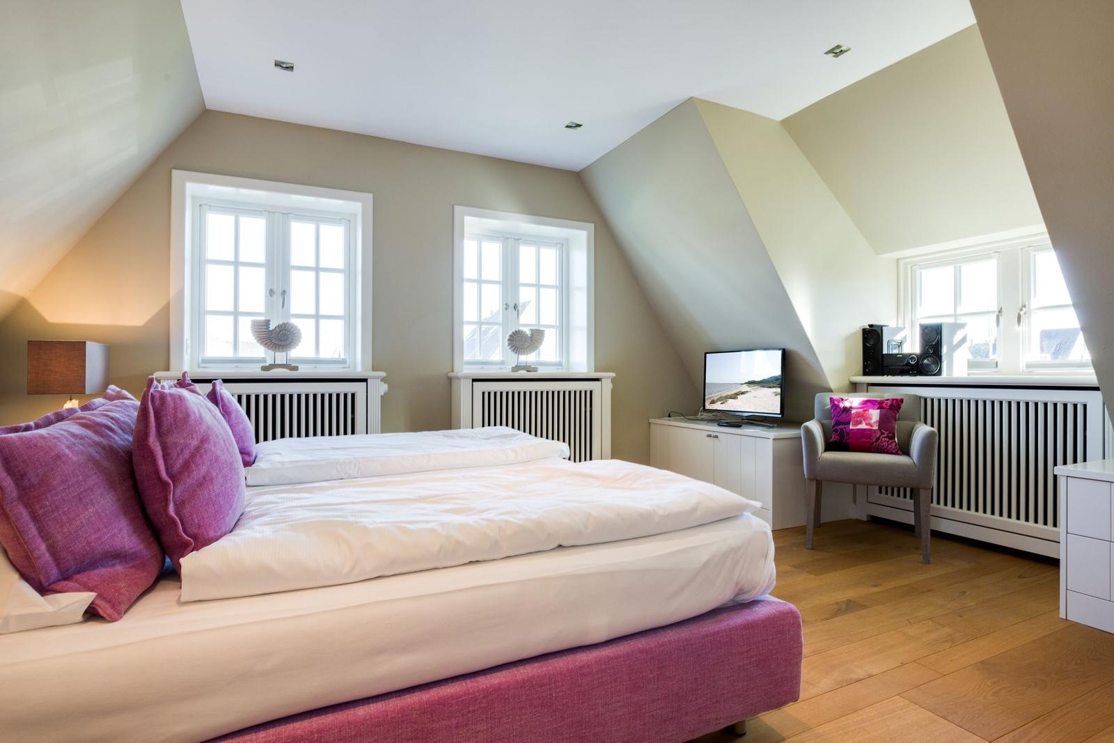 Schlafzimmer mit Doppelbett, zwei Fenstern und Sessel.