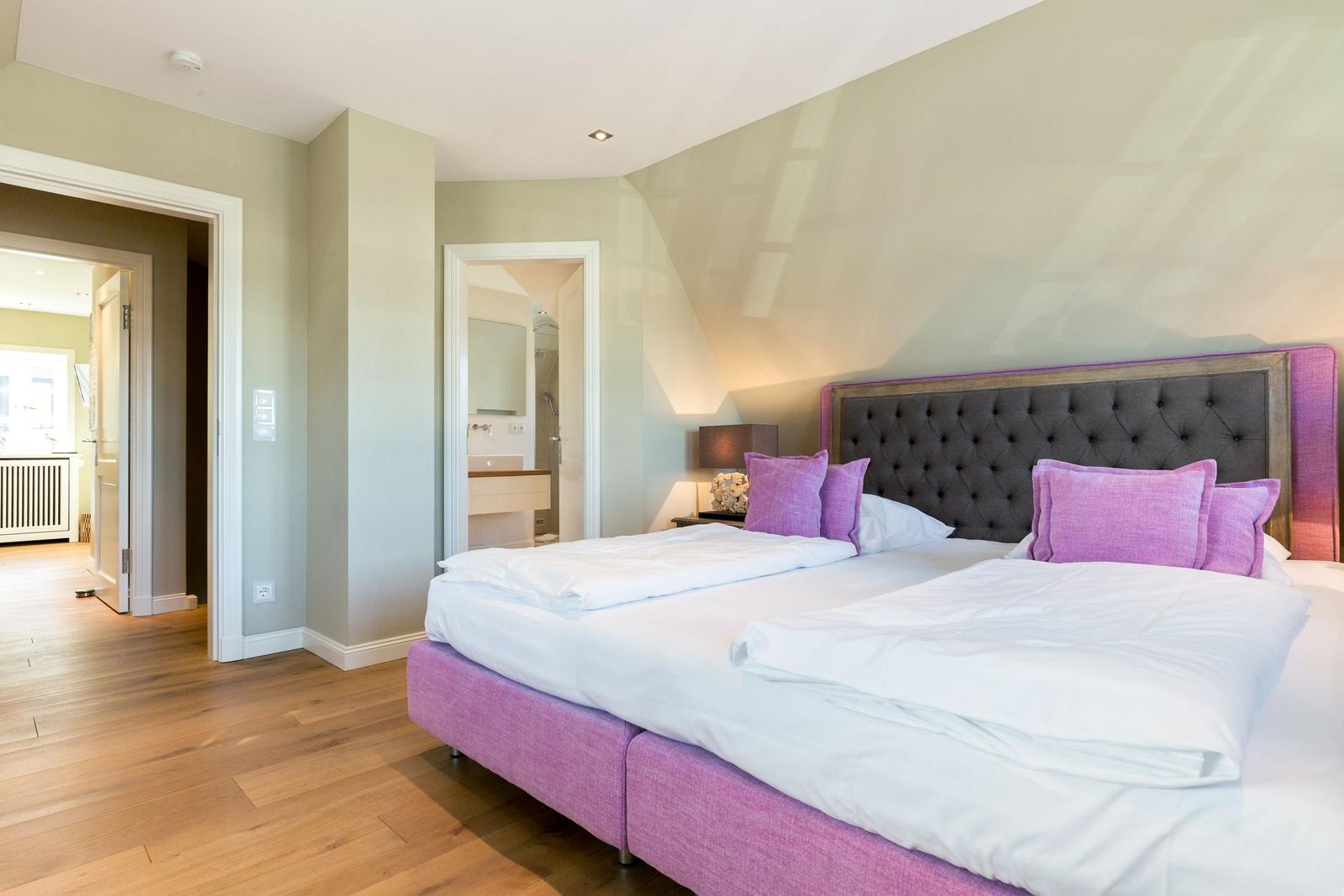Schlafzimmer mit großem Bett, lila Kissen und Holzboden. Blick in Bad und Wohnbereich.