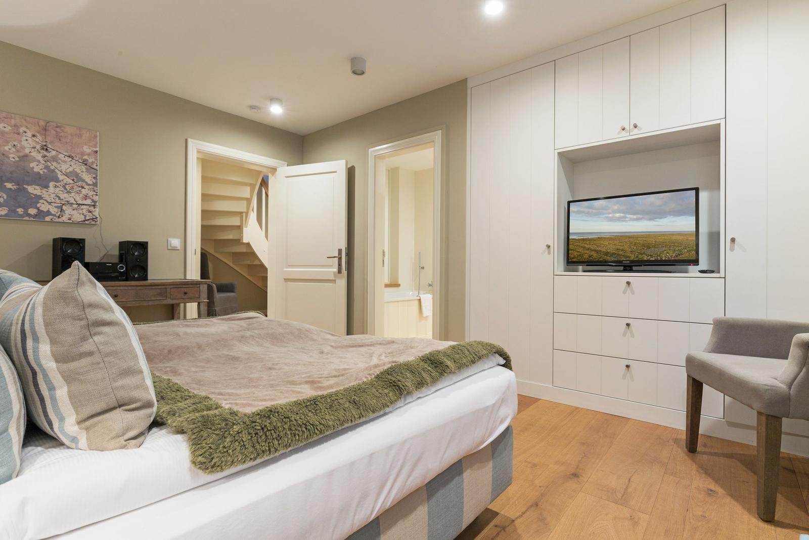 Schlafzimmer mit Bett, TV und Stuhl. Tür führt zur Badewanne und Treppe.