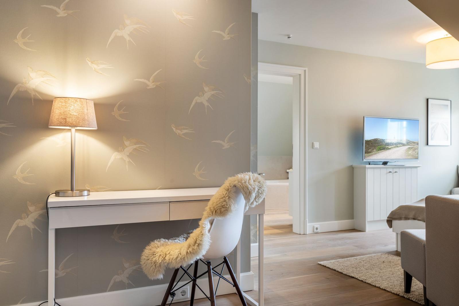 Desk with chair and lamp against bird-patterned wall. TV and seating area in background.