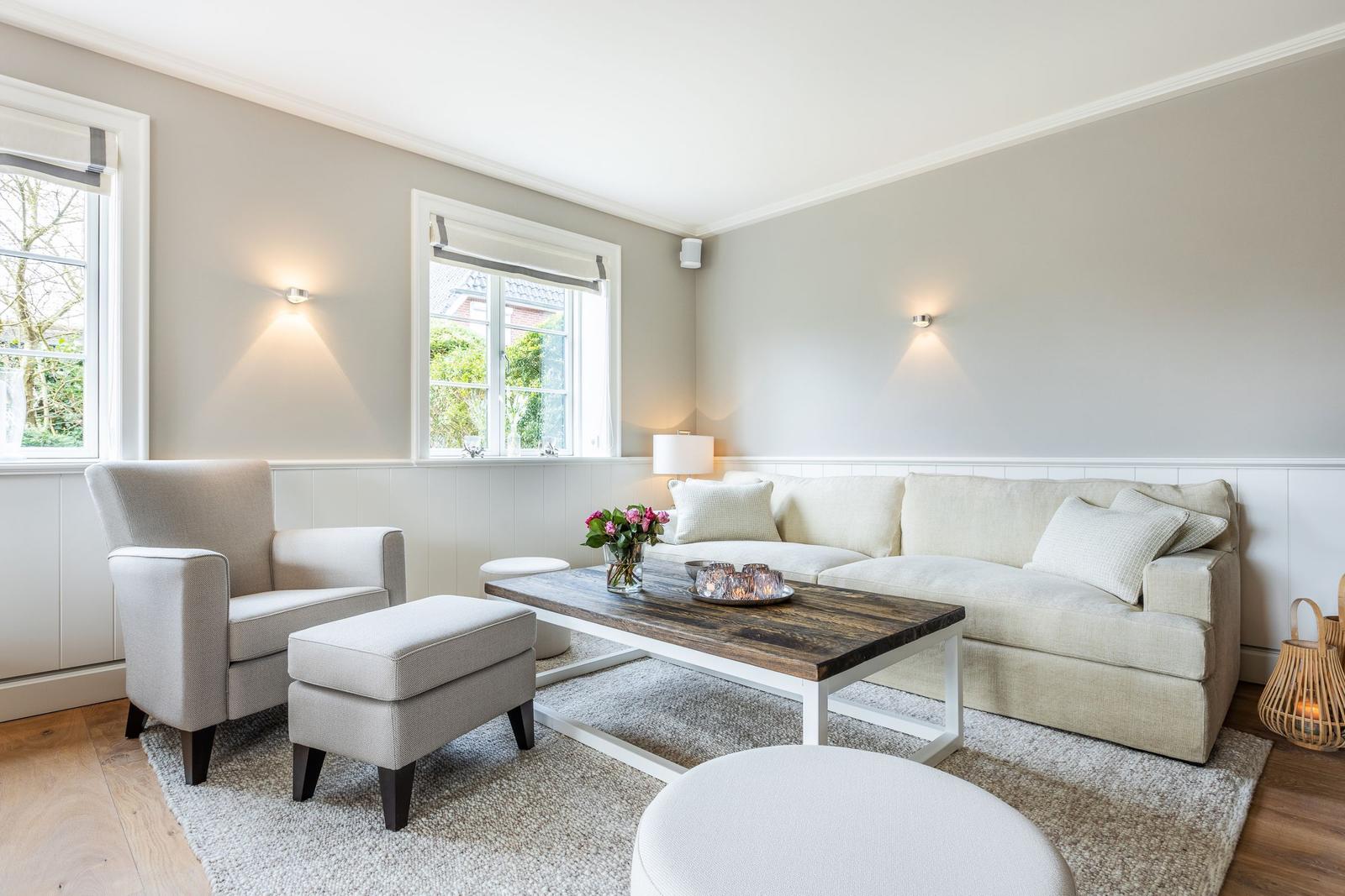 Living room with sofa, armchair, and wooden table. Windows and wall lights.