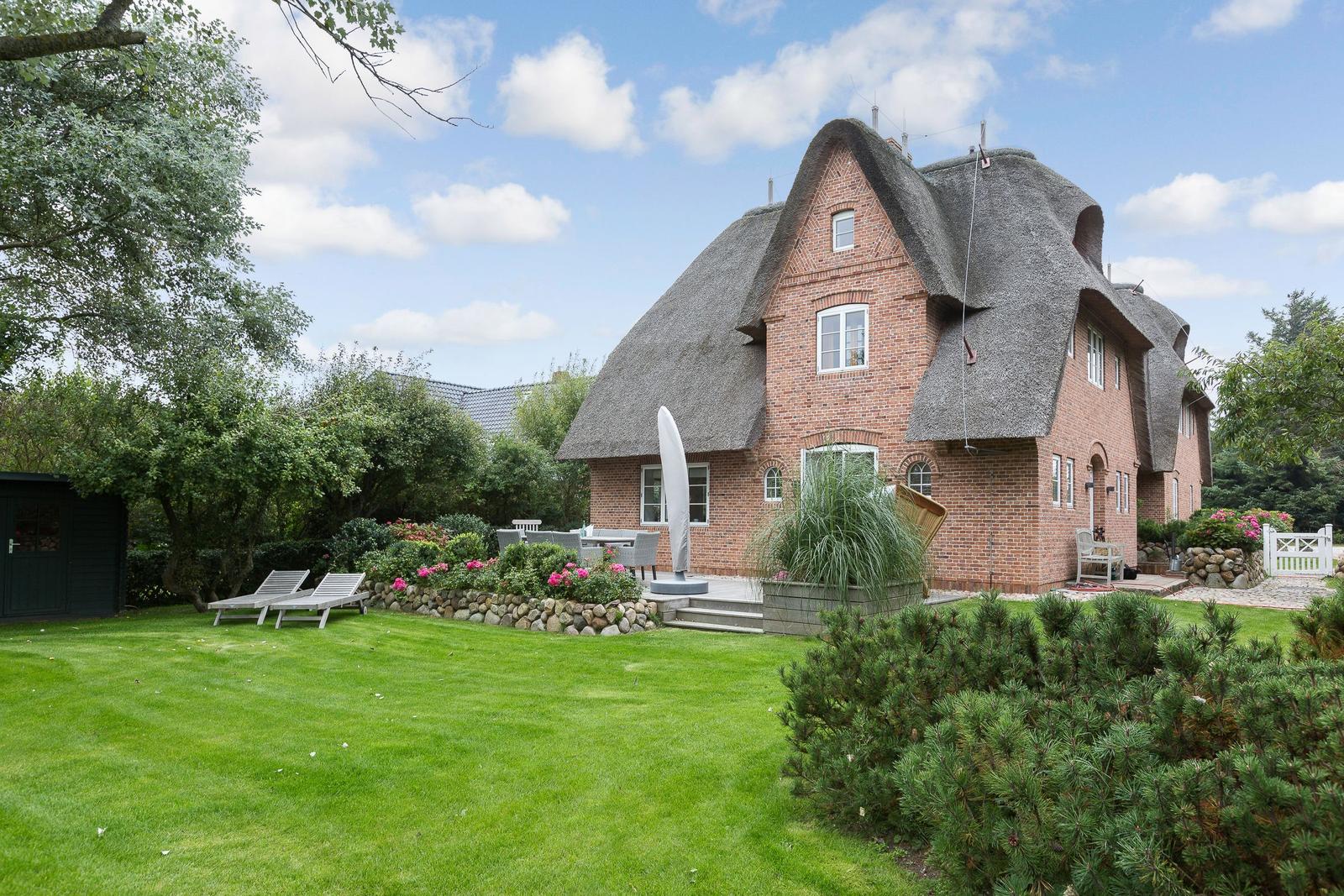 Red brick house with thatched roof, garden with sun loungers and flower beds.