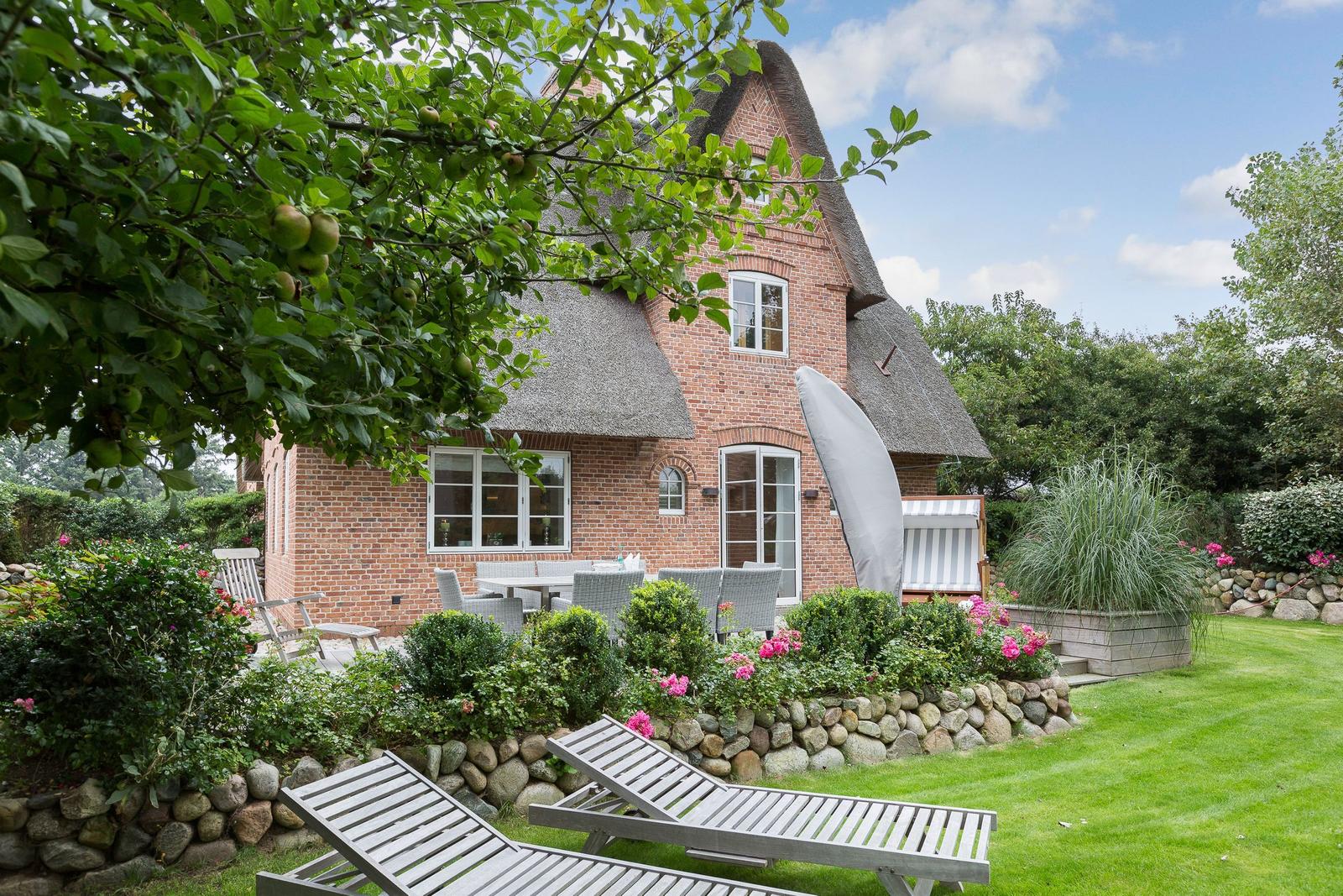 Red brick house with thatched roof, garden with lounge chairs and flower beds.