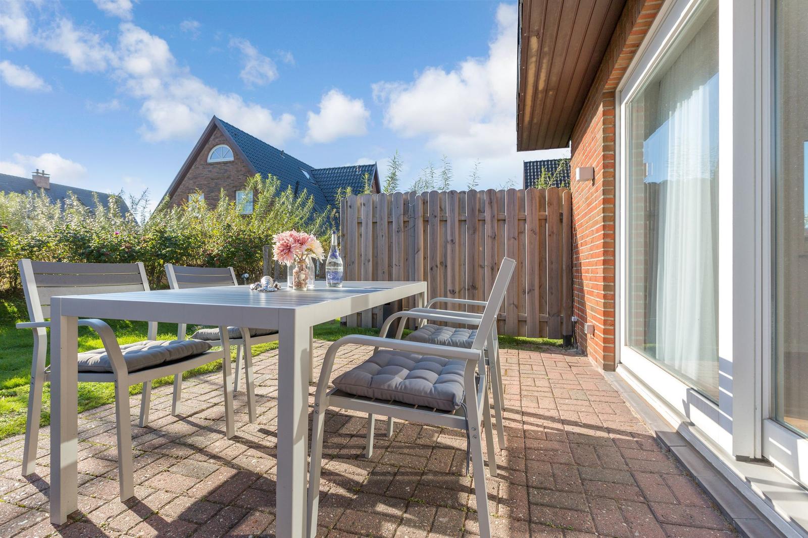 Terrasse mit Tisch und Stühlen, gepflastert, mit Blick auf einen Garten und Zaun.