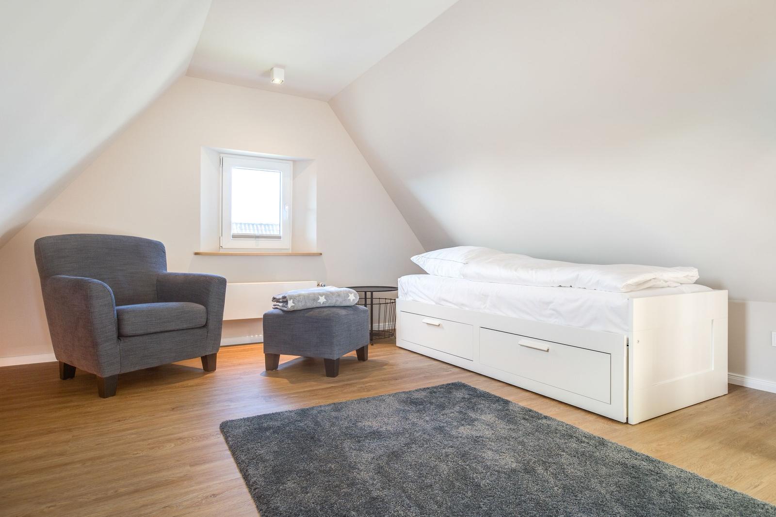 Schlafzimmer mit Dachschrägen, weißem Bett und grauer Sitzgelegenheit.