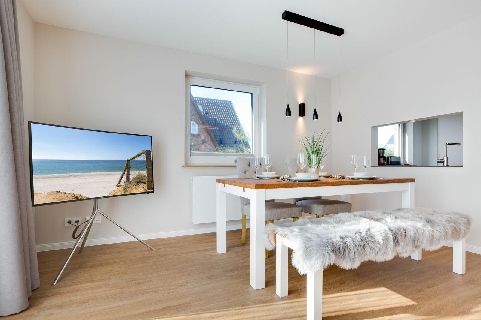 Essbereich mit Tisch, Stühlen und Fernseher. Fenster zeigt Haus und Strand.