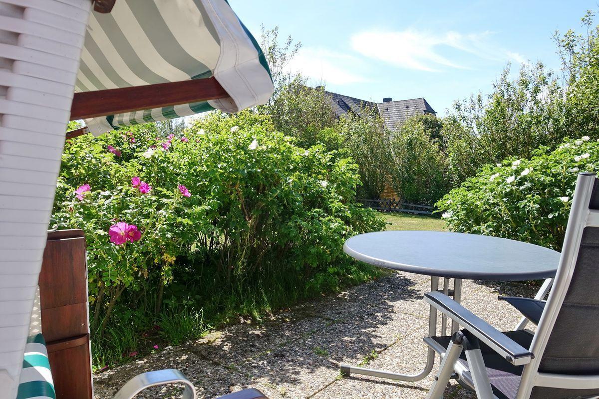 Terrace with table, chairs, and sunshade. Background: green hedge and roses.