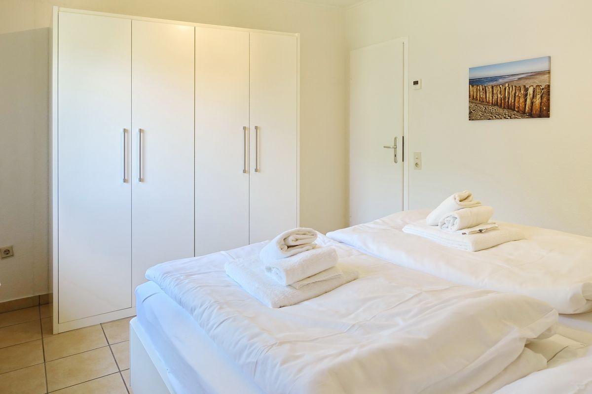 Two beds with white sheets and folded towels. Background: white wardrobe and wall picture.