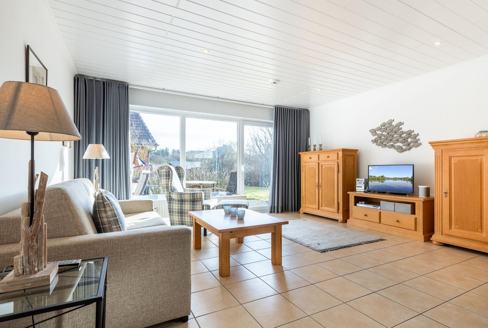Living room with sofa, coffee table, and TV. Large windows with garden view.
