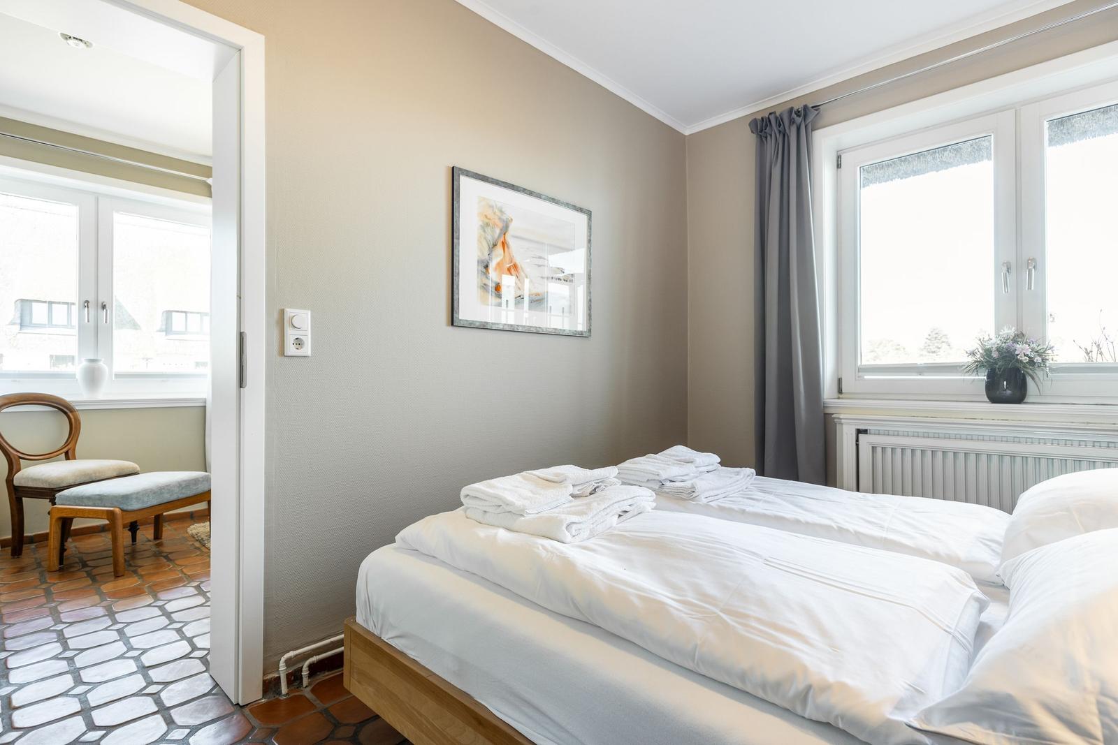 Schlafzimmer mit Bett, Fenster und Bild an der Wand. Durchgang zu einem Raum mit Stuhl.