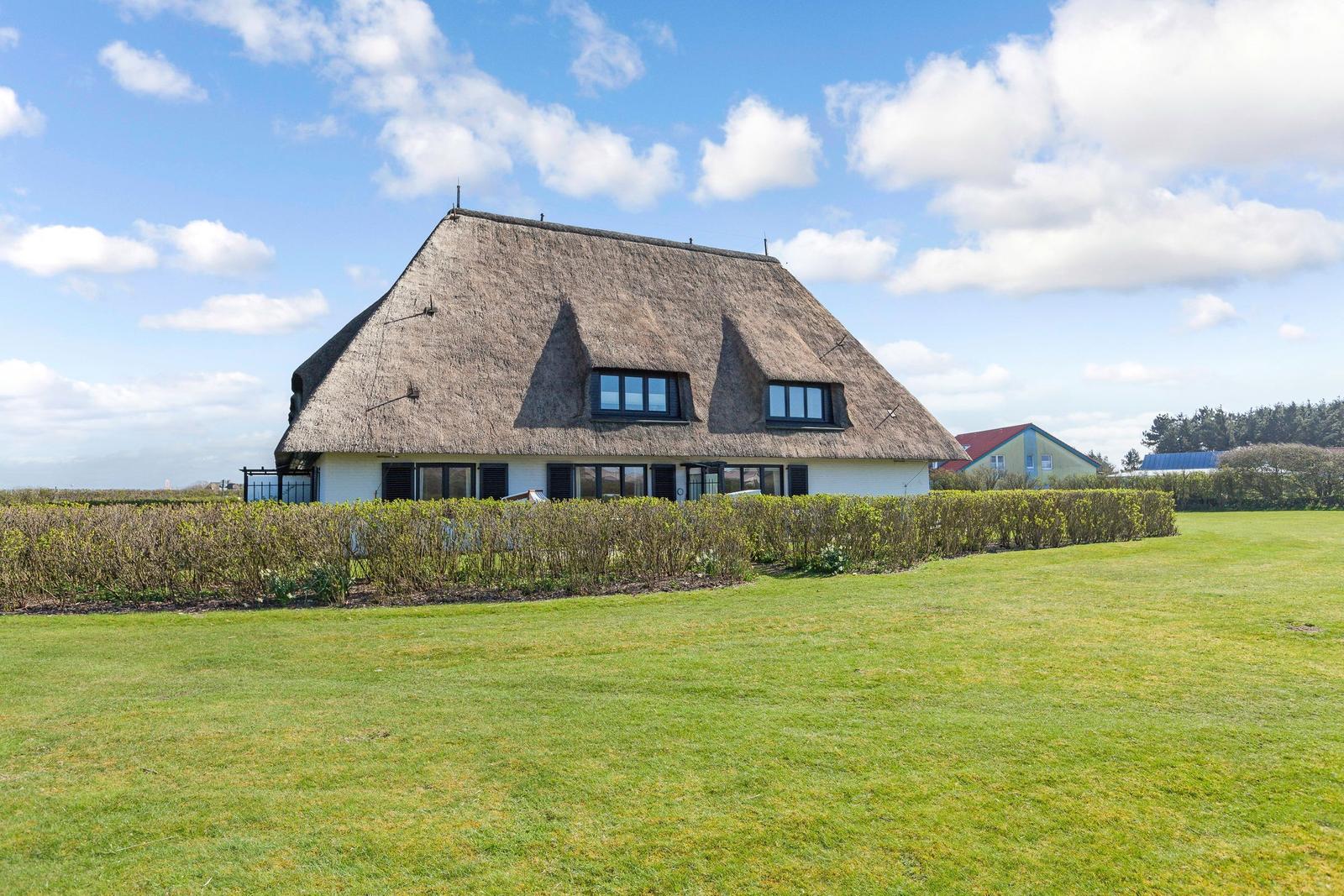 Haus mit Strohdach, umgeben von Gras und Hecken unter blauem Himmel.